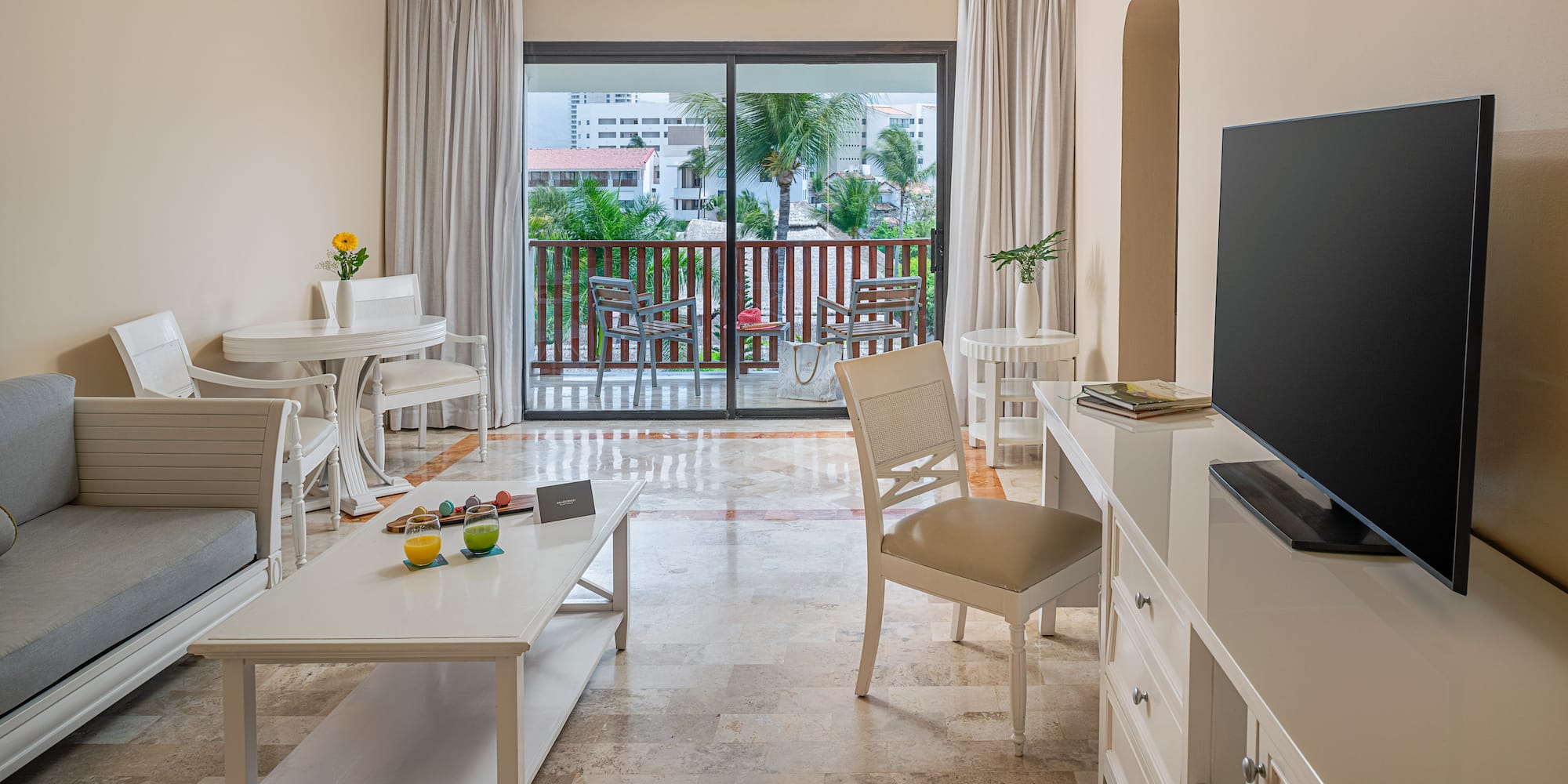 Bright hotel suite living room with balcony overlooking tropical resort.