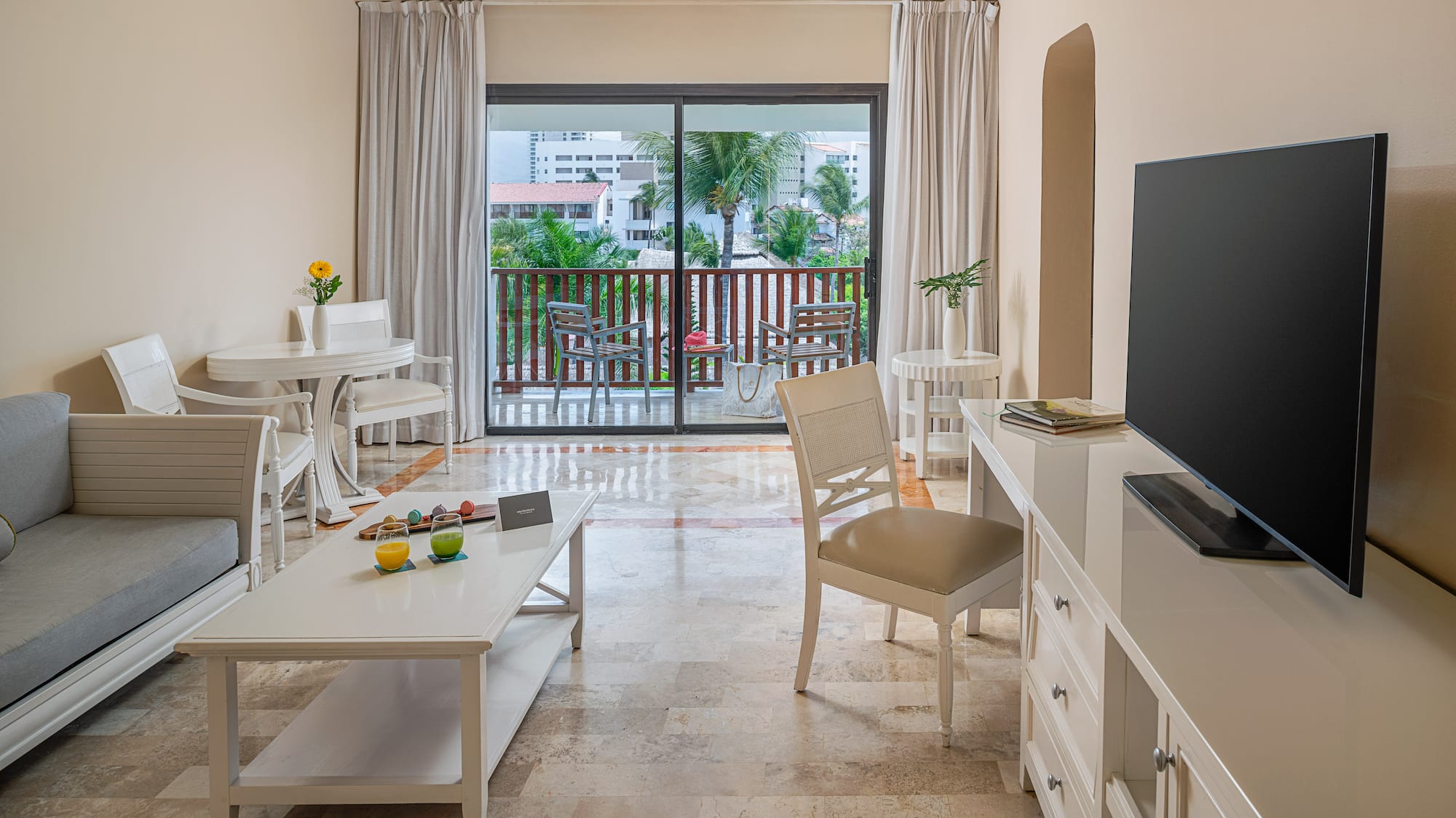 Spacious hotel suite living area with balcony, white furniture, tropical view.