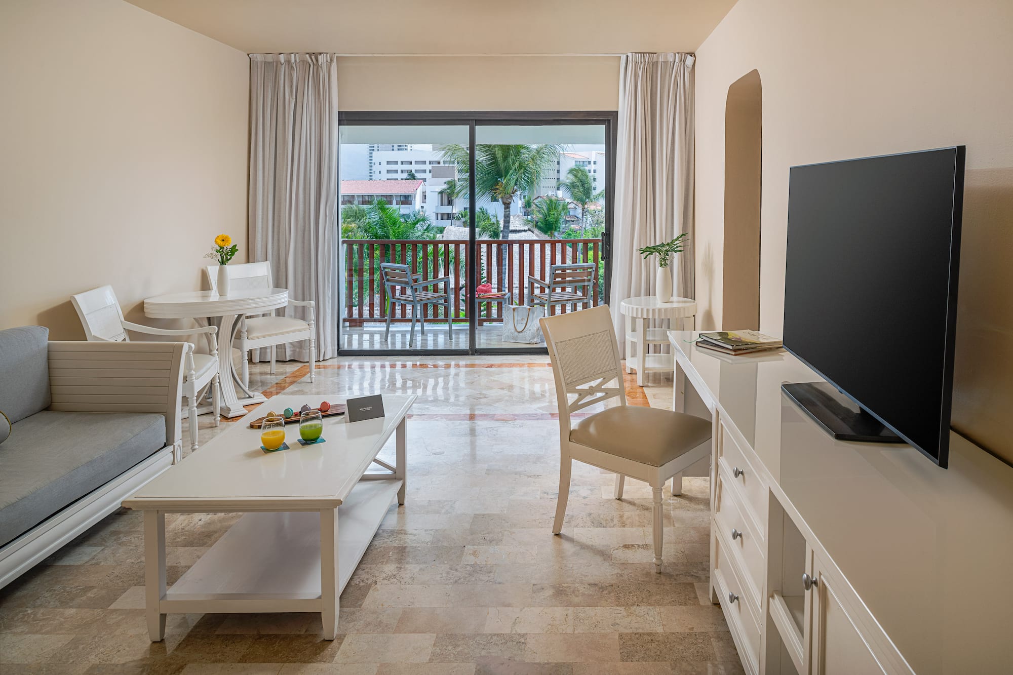 Bright hotel suite living room with balcony overlooking tropical resort.