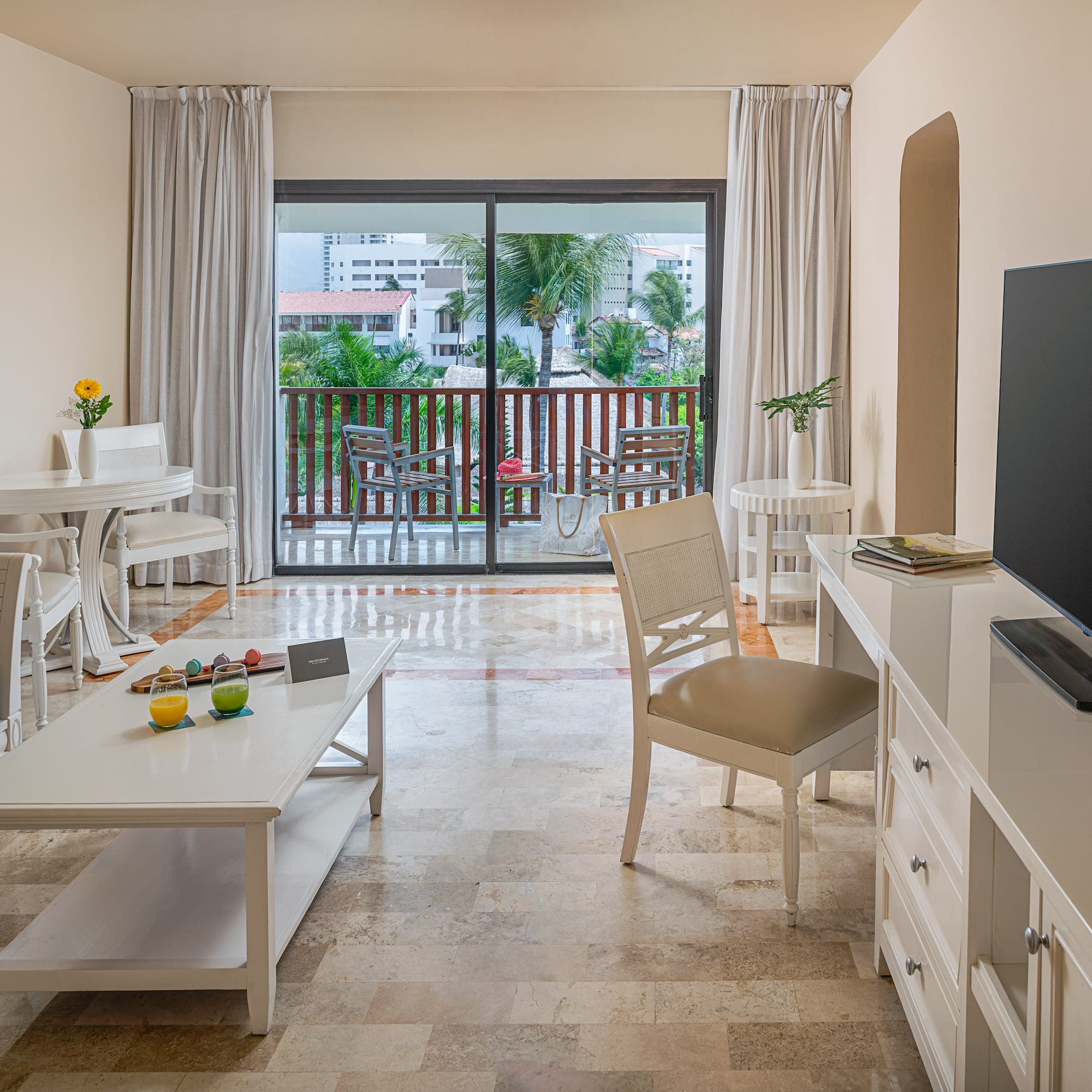 Spacious hotel suite living area with balcony, white furniture, tropical view.