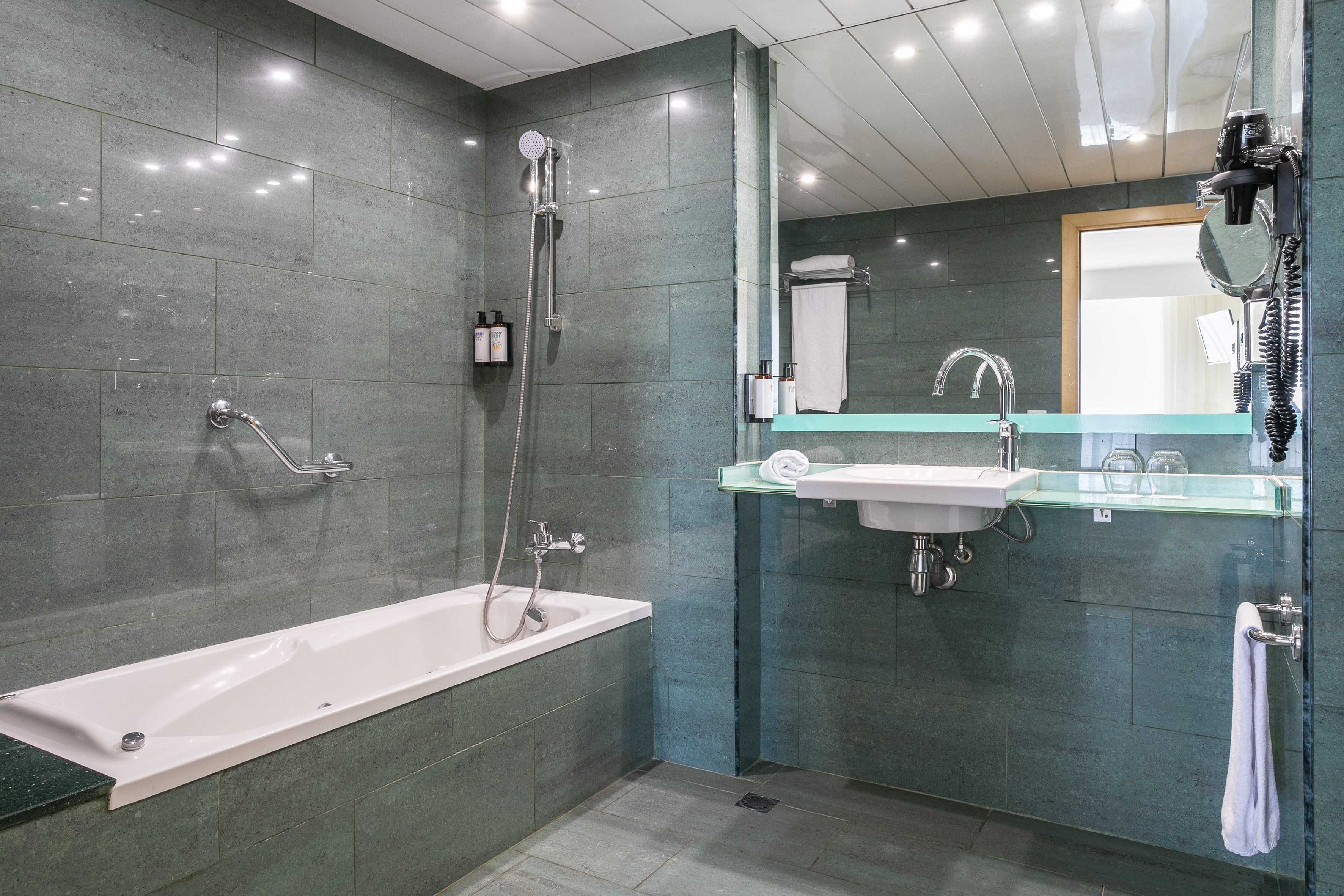 Hotel bathroom with green tiled walls, bathtub, shower, sink, and large mirror.