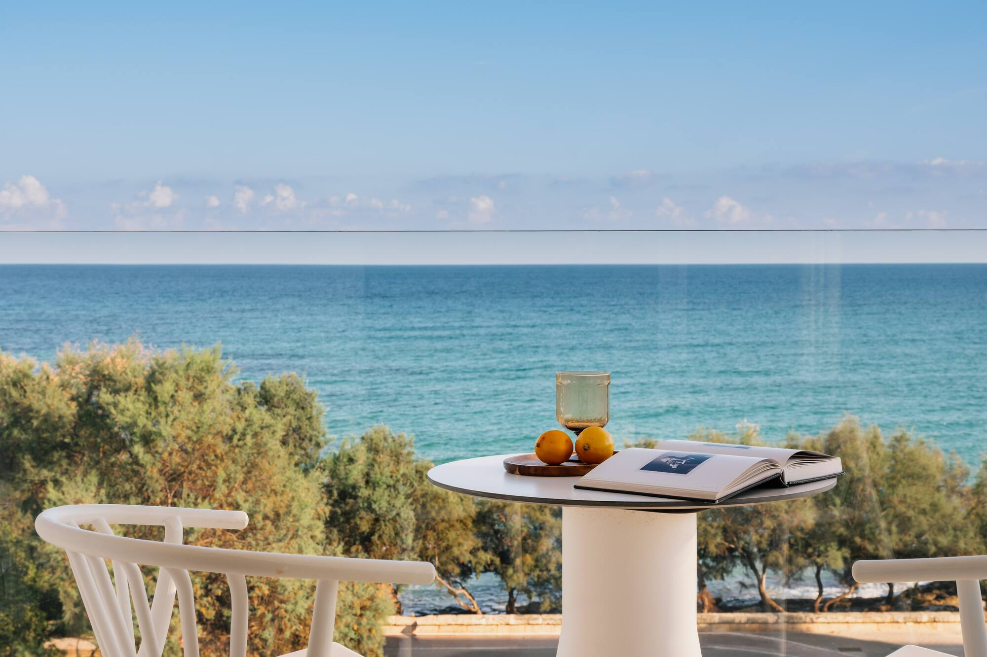 Balcón con vista al mar, mesa, libro y naranjas bajo cielo azul.