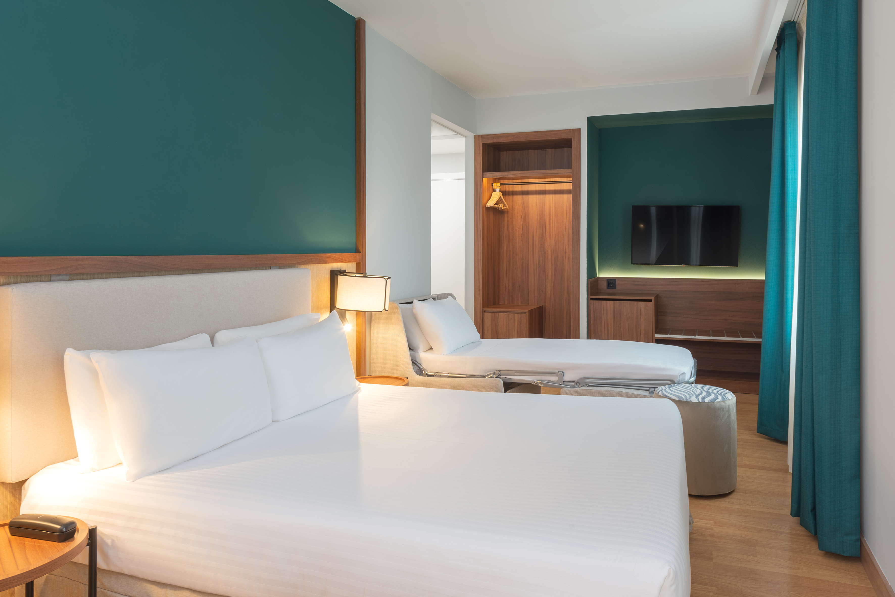 Modern hotel bedroom with large bed, sofa bed, teal walls, wood furniture, and TV.