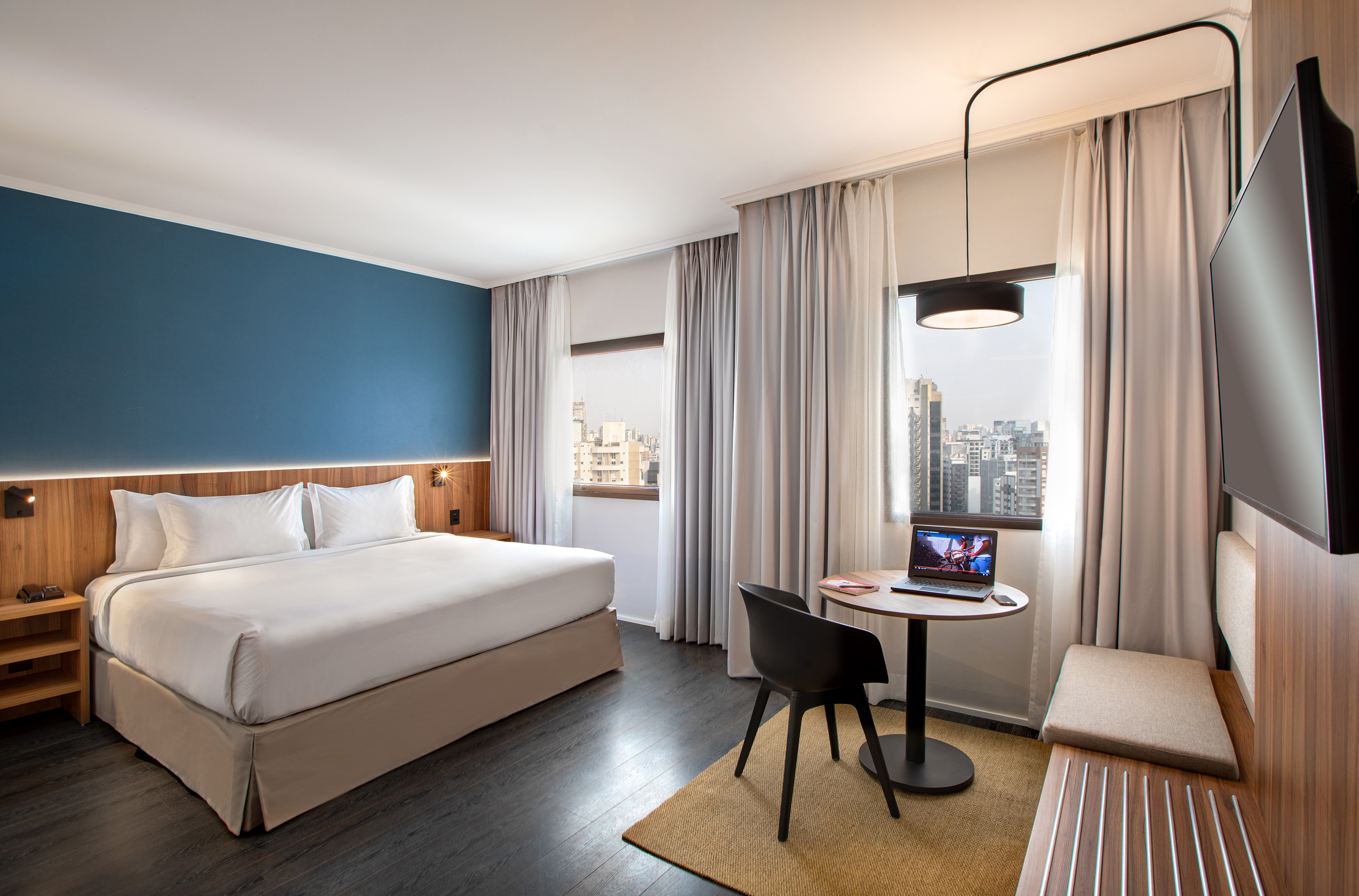 Modern hotel room with king bed, blue wall, city view, and laptop workspace.