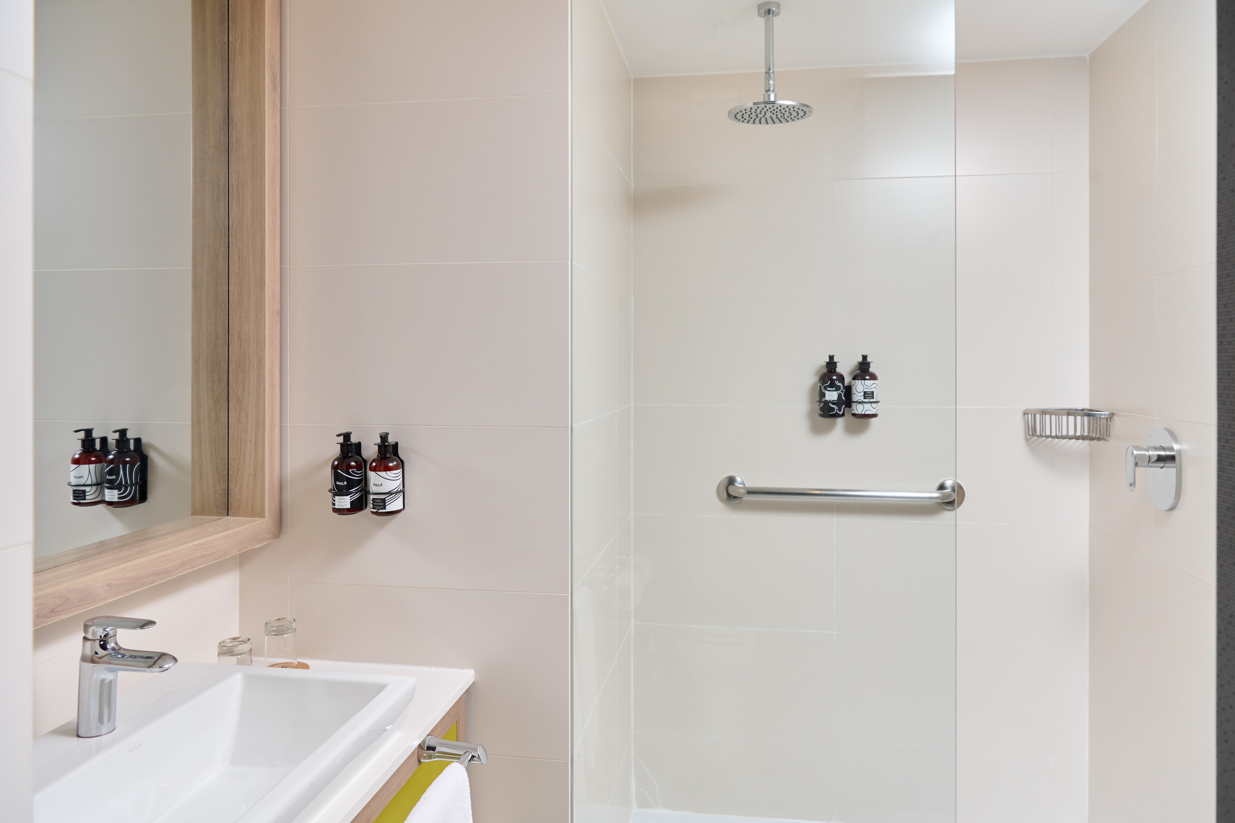 Modern hotel bathroom with sink, mirror, rainfall shower, and grab bar.