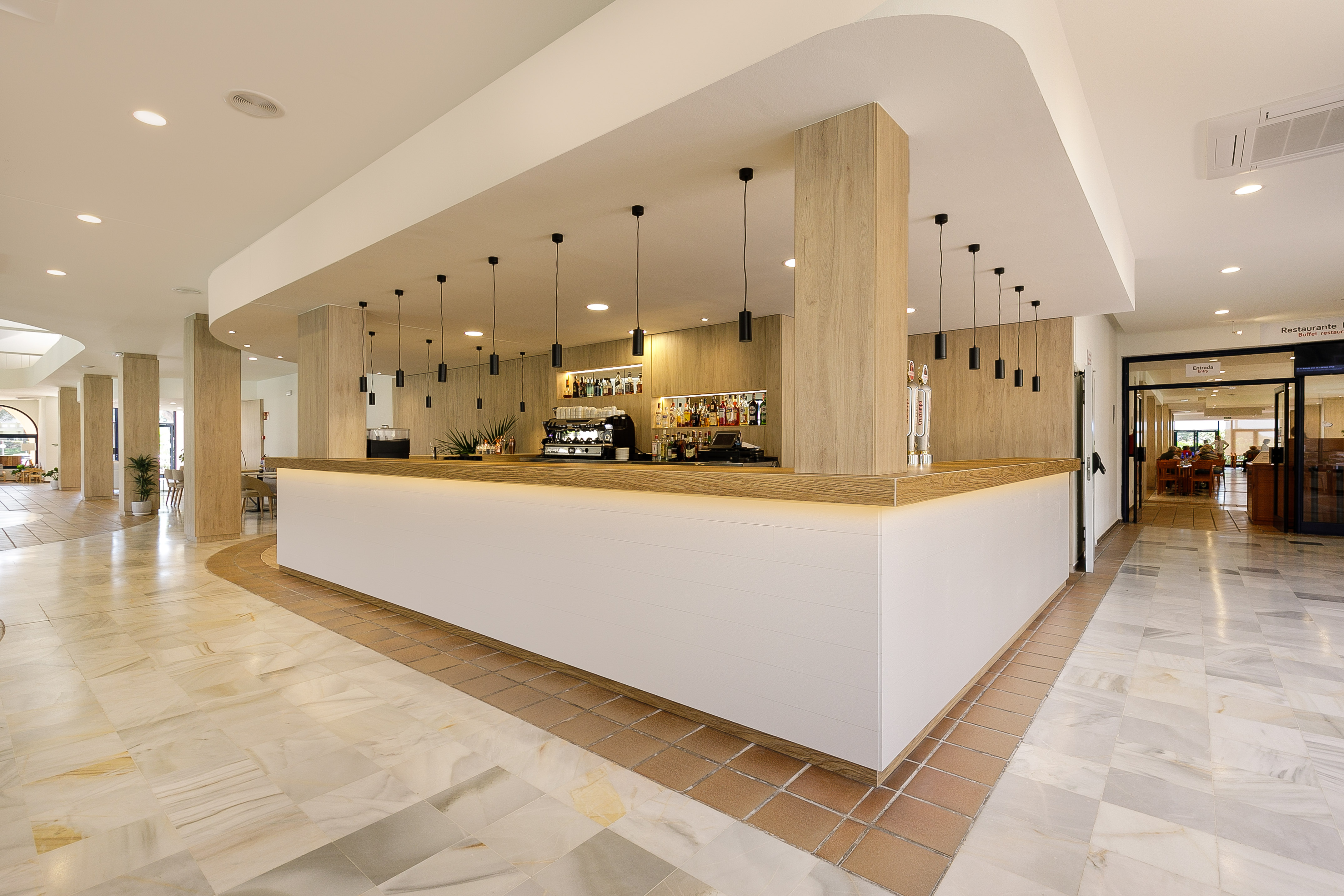 Modern hotel bar with white and wood counter, pendant lights, and tiled floor.