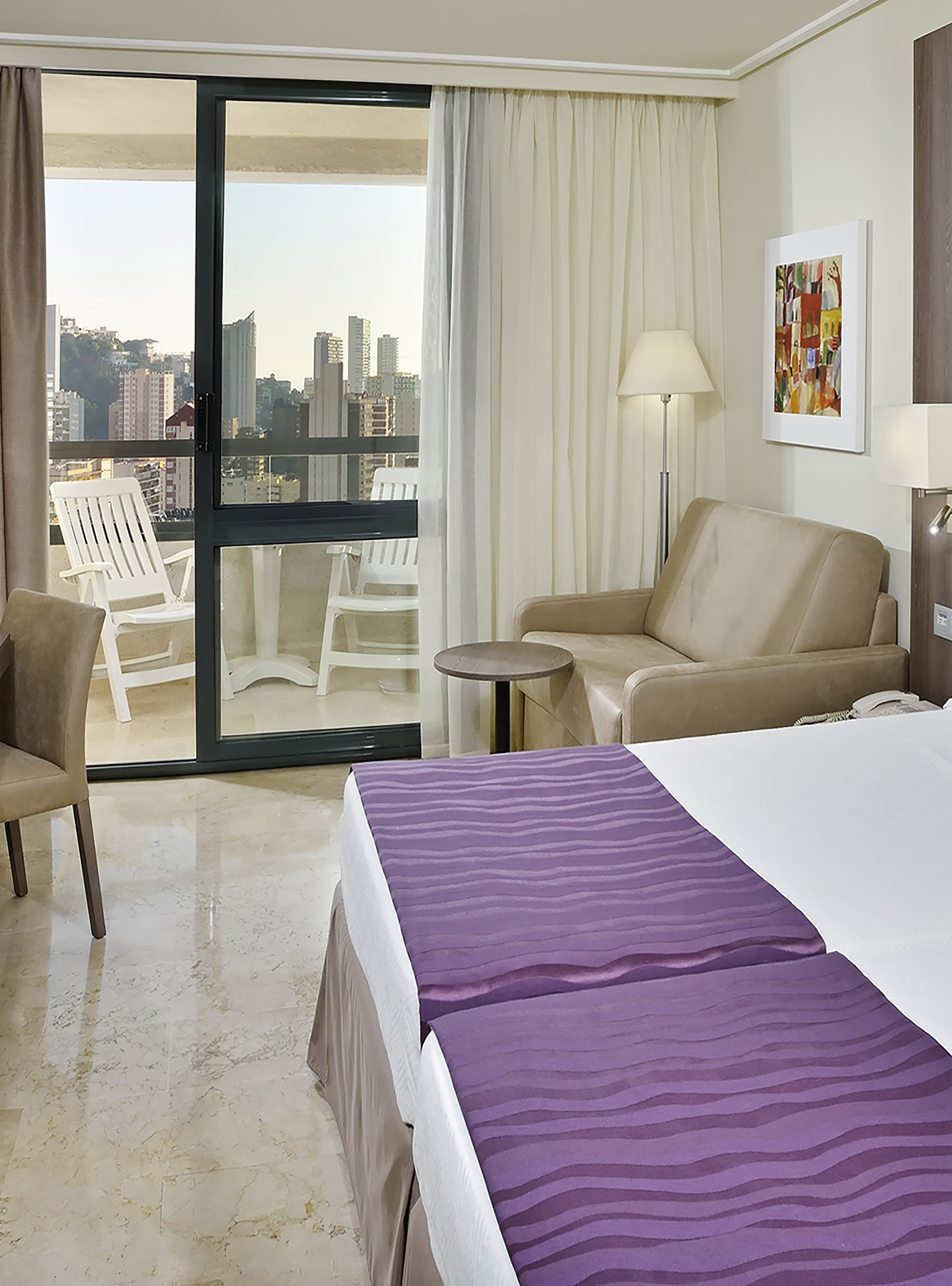 Hotel room with city view balcony, bed with purple runners, and sofa.