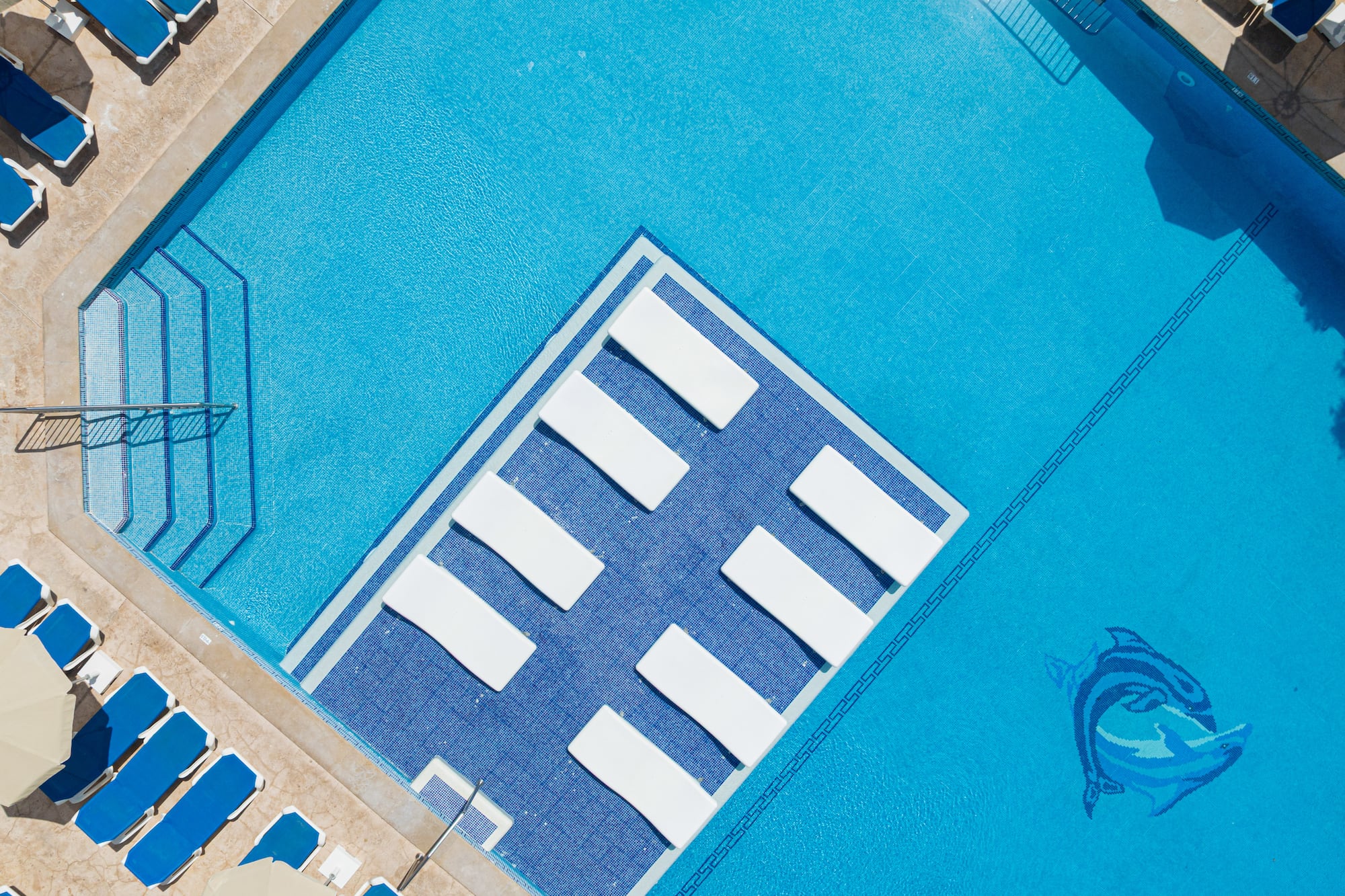 Aerial view: bright blue resort swimming pool with in-water loungers, sunbeds.