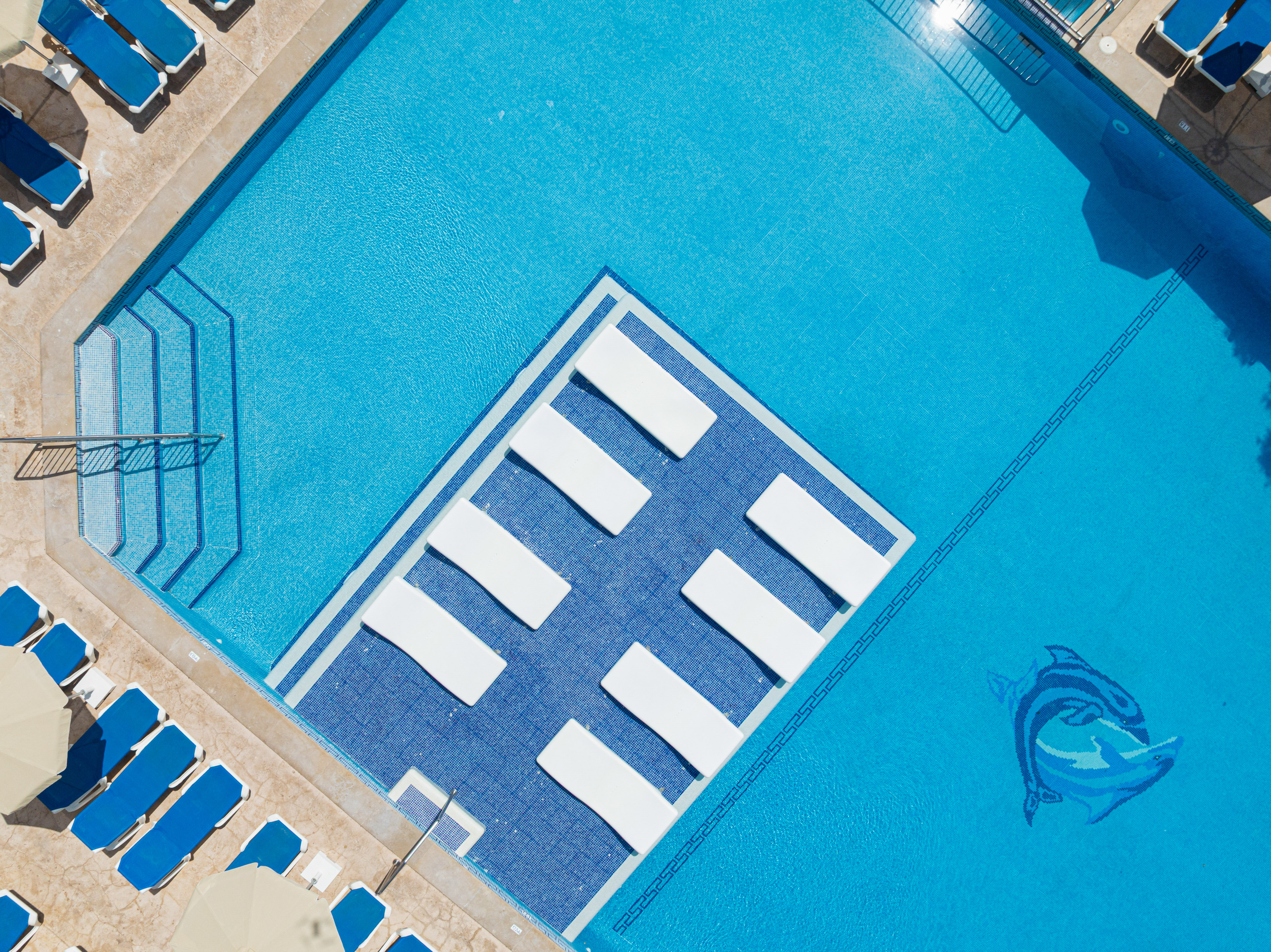 Aerial view: bright blue resort swimming pool with in-water loungers, sunbeds.