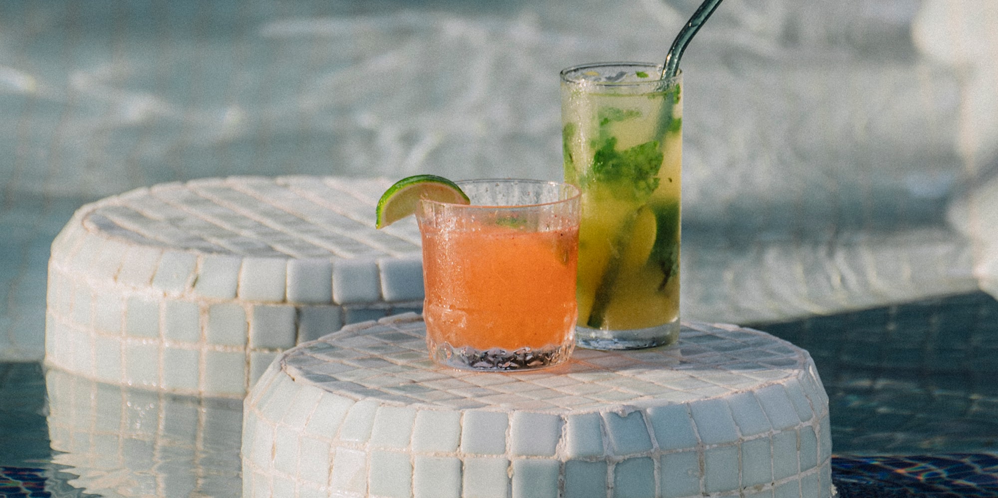 Orange and green cocktails on tiled tables in a swimming pool.