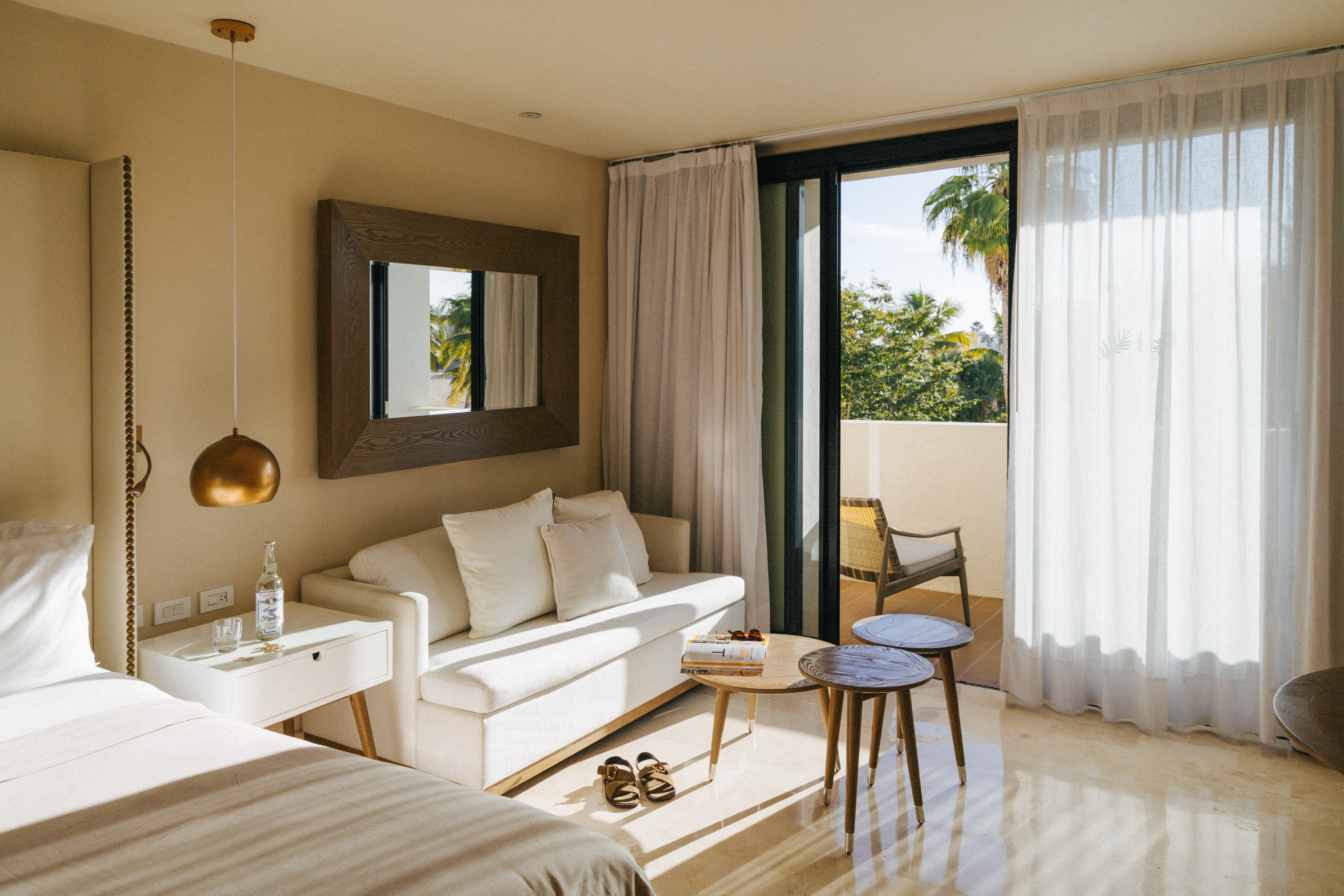 Sunny hotel suite with bed, white sofa, balcony, and tropical view.