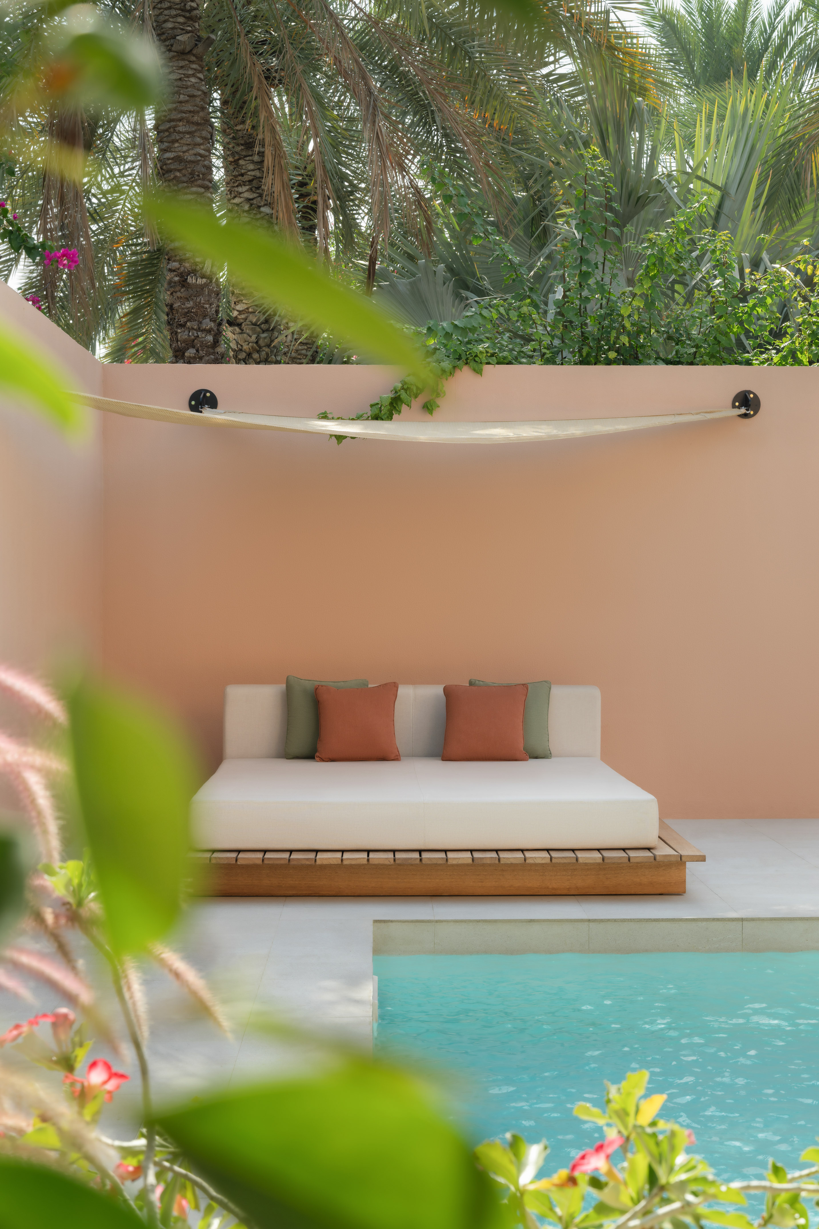 Daybed with terracotta pillows beside a turquoise pool, peach wall, and palms.