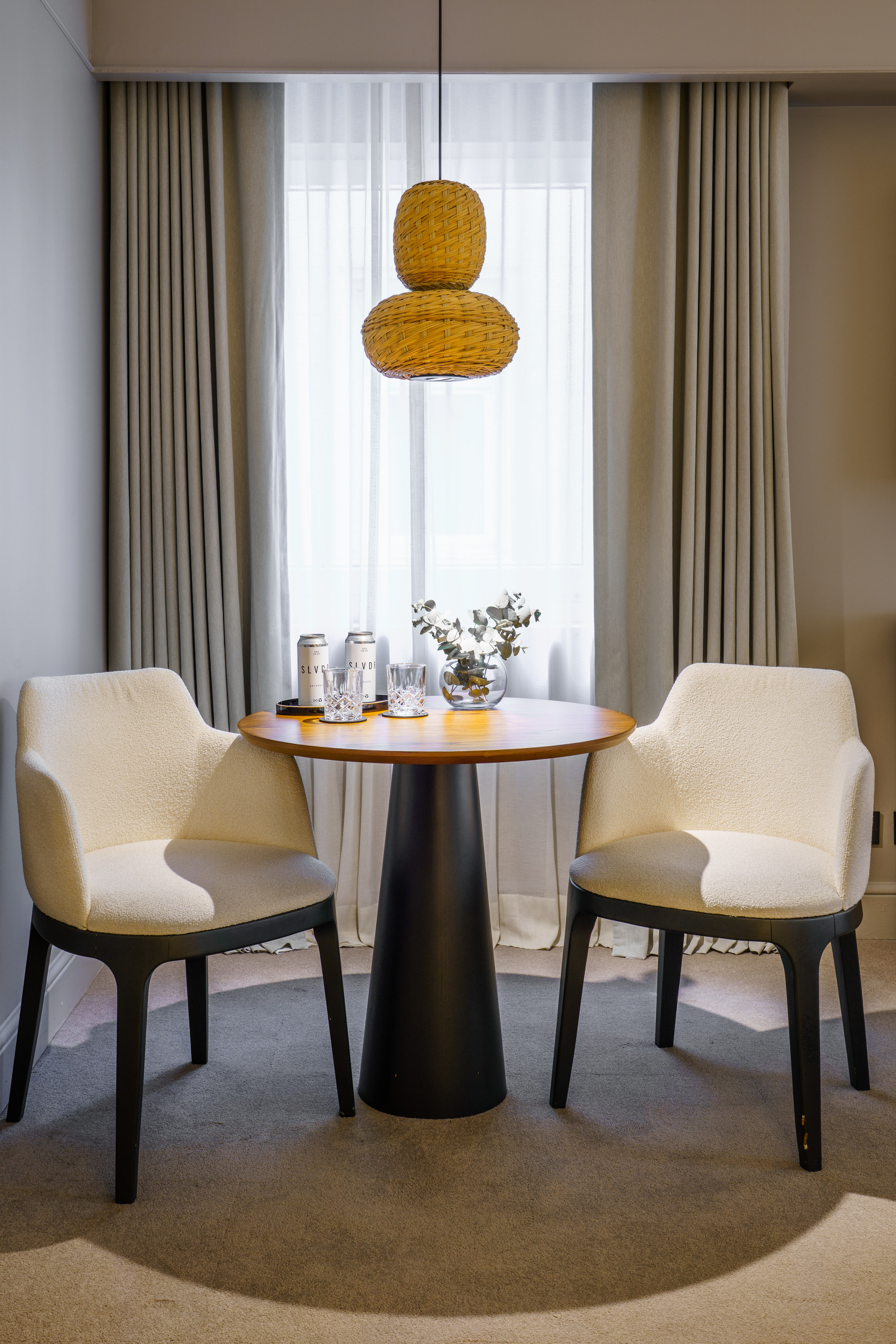 Modern hotel dining area with round table, two cream chairs, rattan lamp.