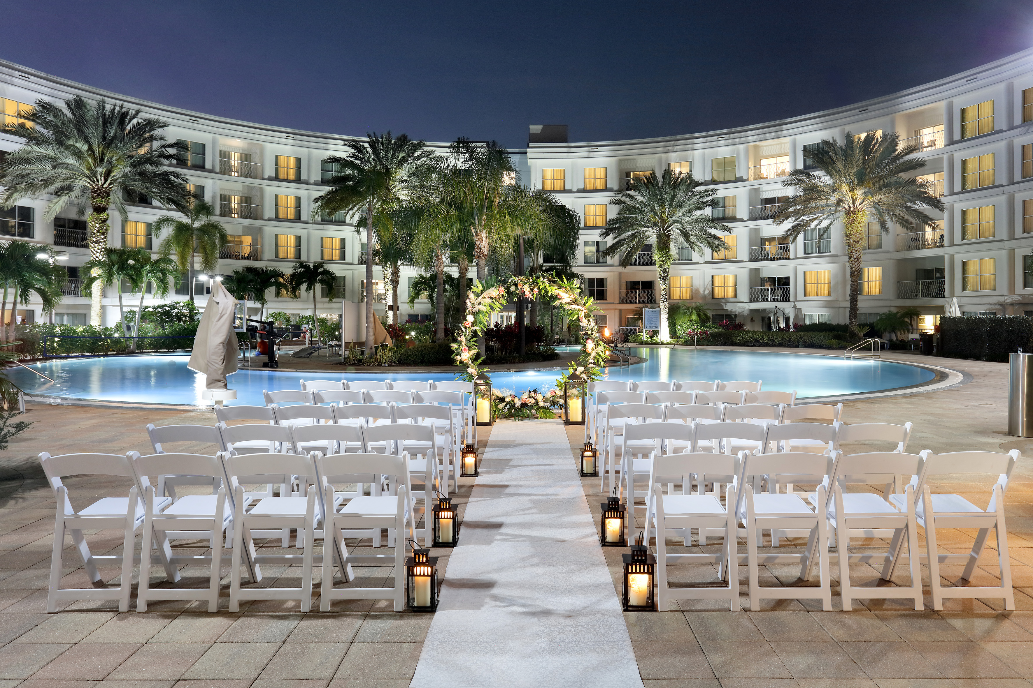 Tropical night wedding ceremony by hotel pool, floral arch, white chairs. - Pool Side (outdoors) - Meliá Orlando Celebration, Orlando