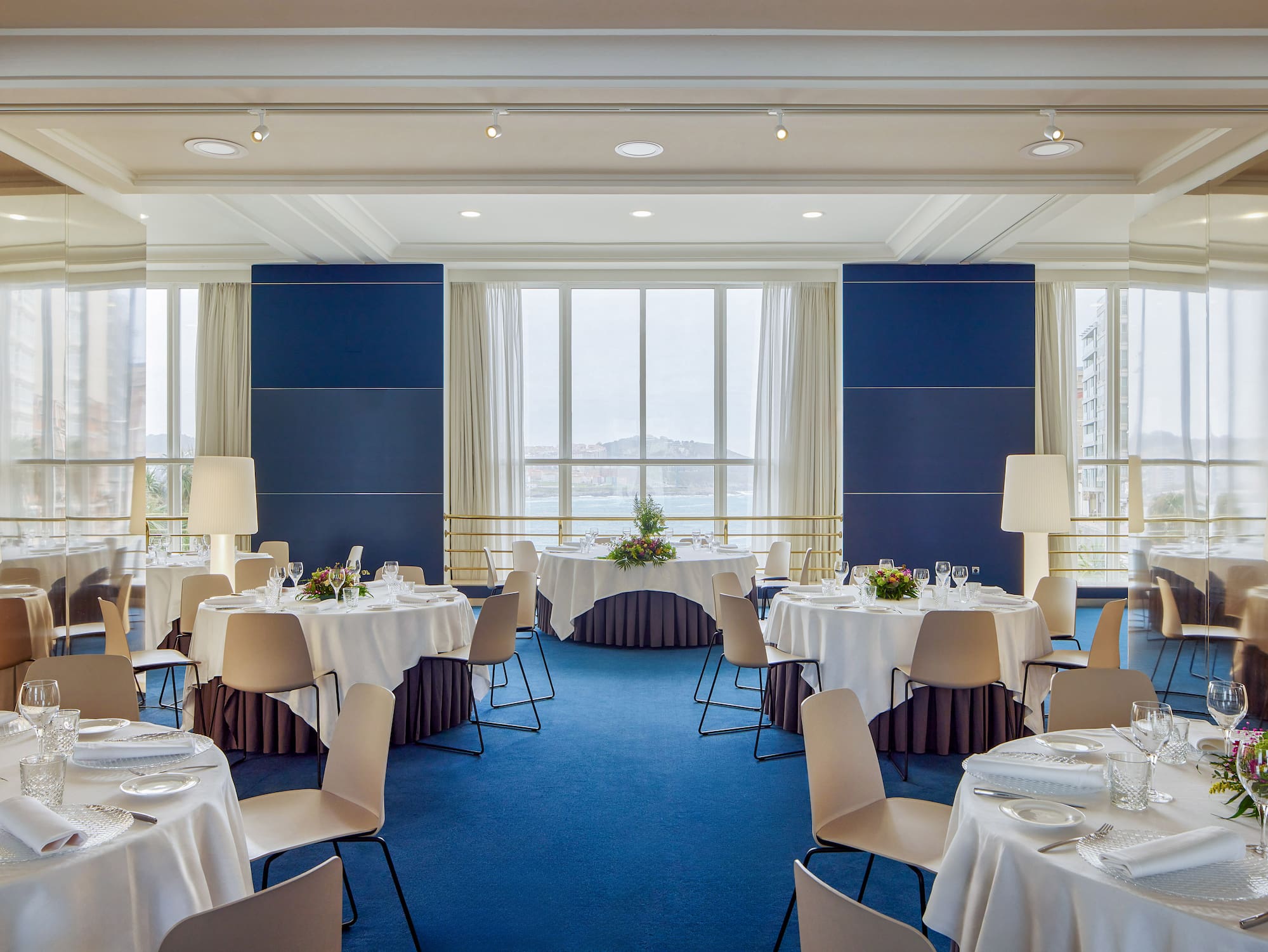 Salón de eventos elegante con mesas redondas, sillas modernas y vistas al mar.