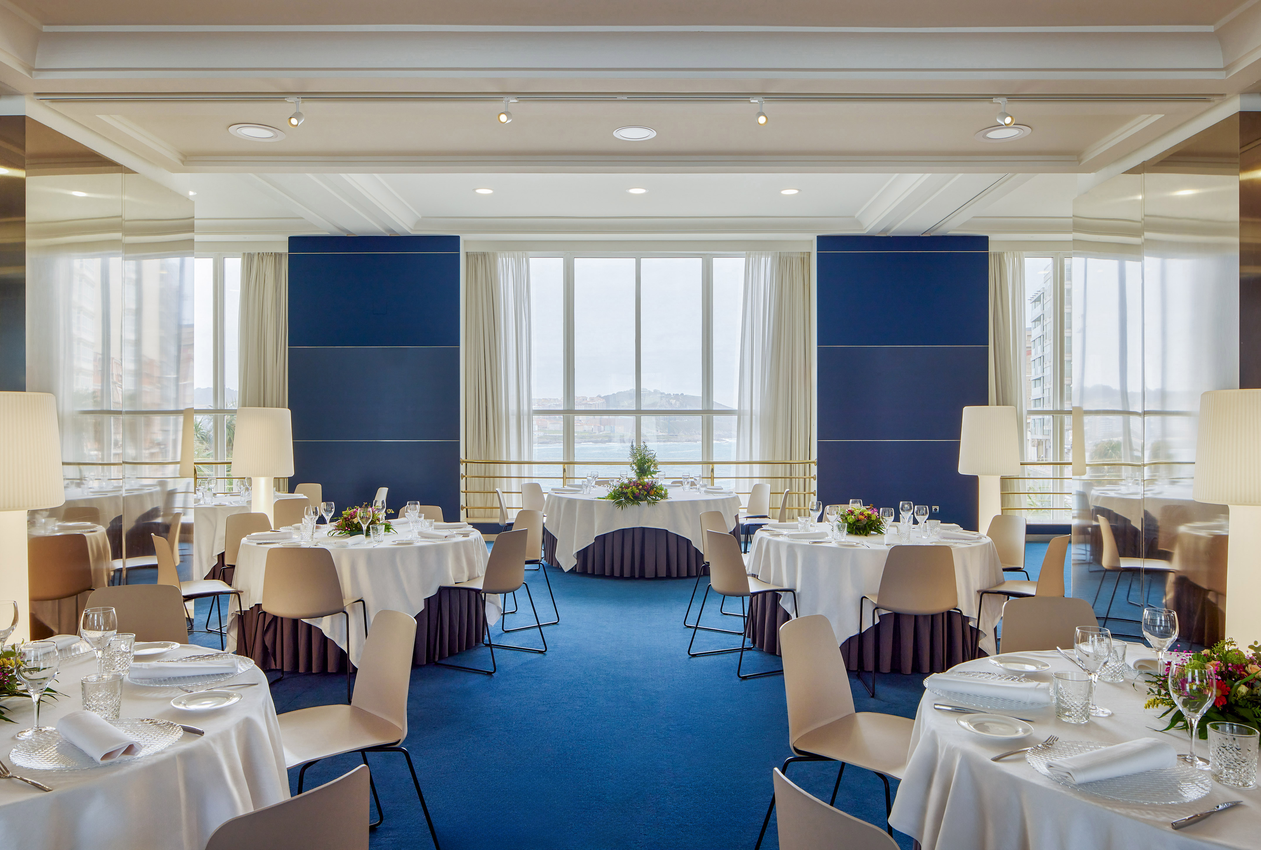 Salón de eventos elegante con mesas redondas, sillas modernas y vistas al mar.