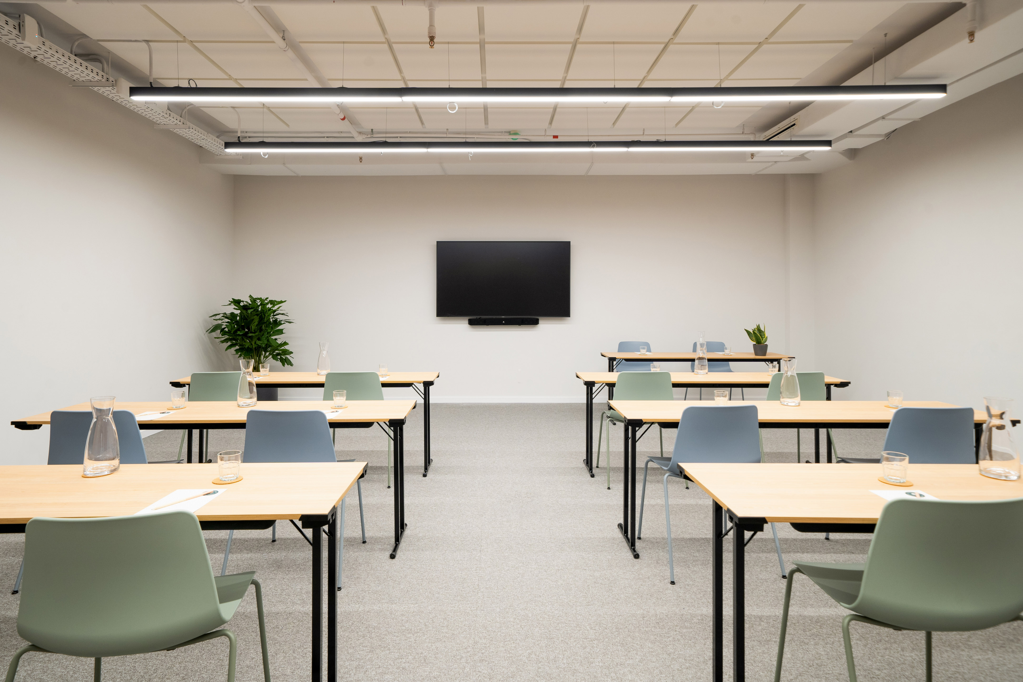 Bright, modern meeting room with tables, chairs, and a large wall TV.