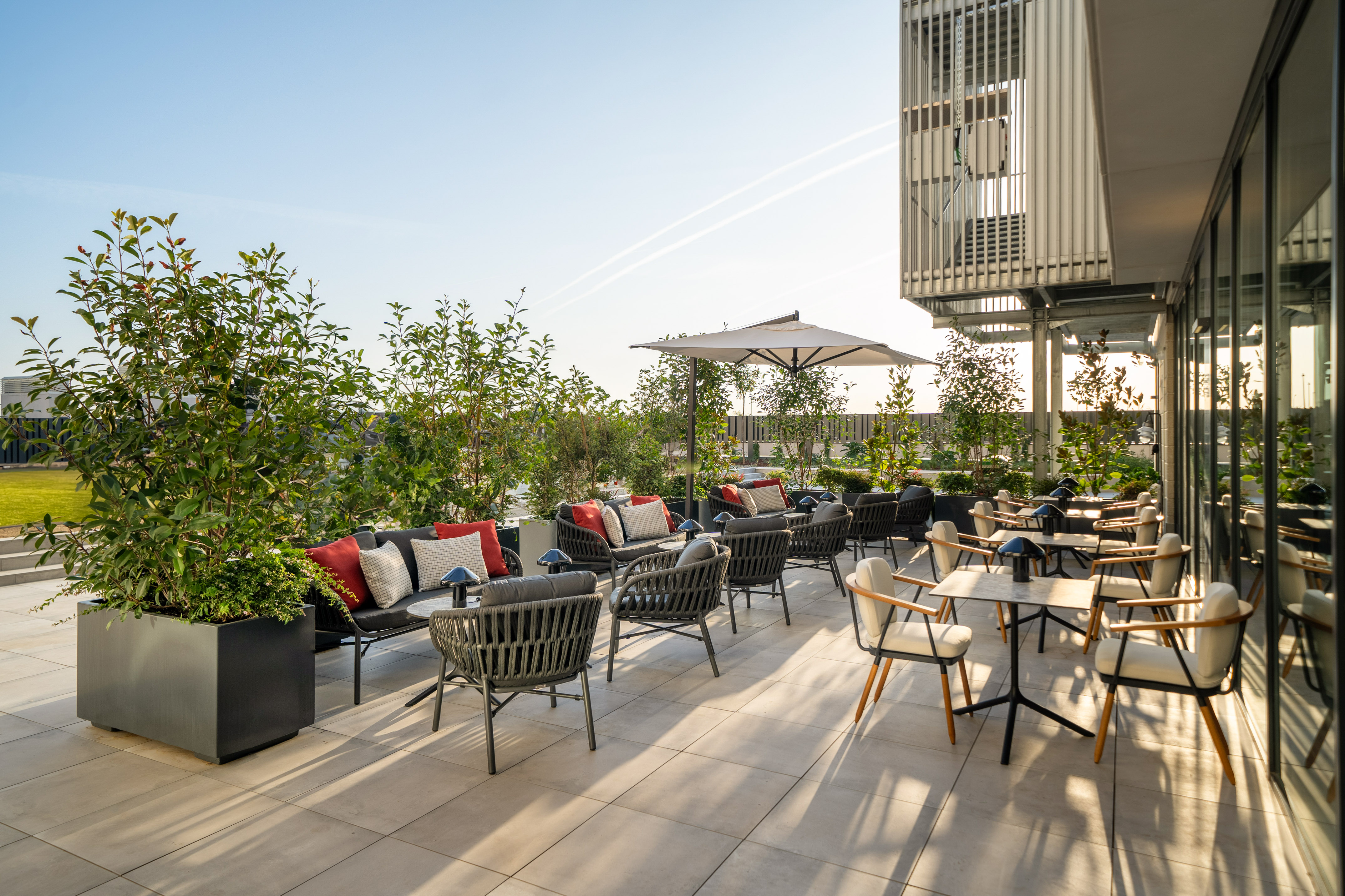 Sunny outdoor patio with modern lounge seating, tables, umbrella, and lush greenery.