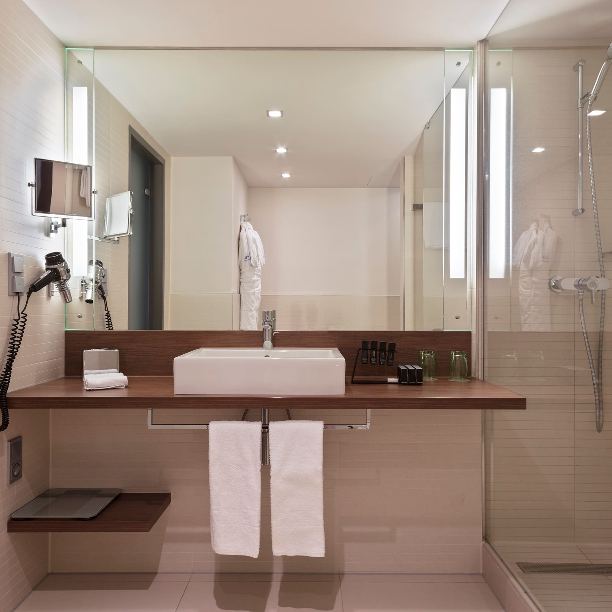 Modern hotel bathroom with white sink, wooden vanity, large mirror, and glass shower.