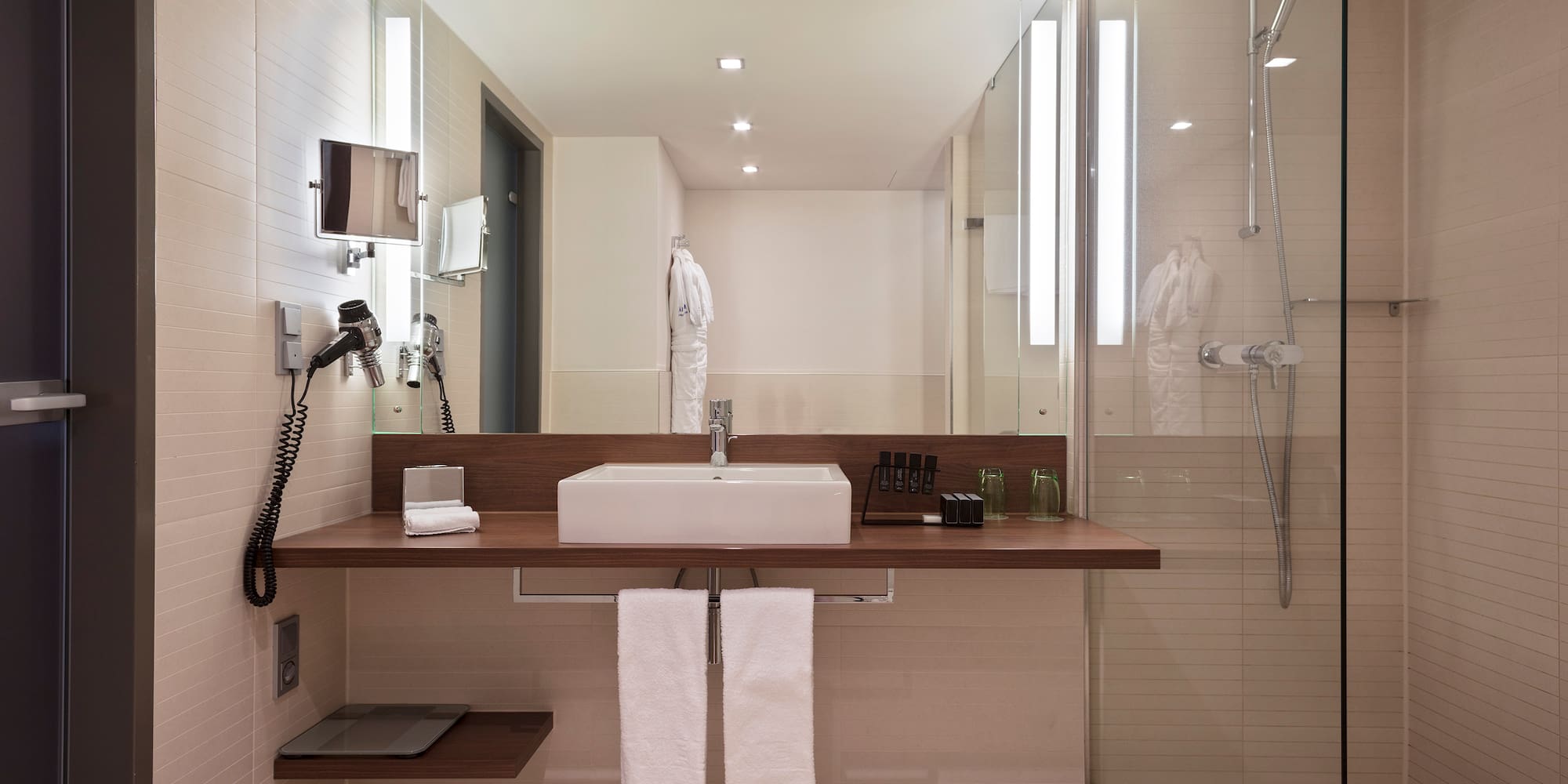 Modern hotel bathroom with white sink, wooden vanity, large mirror, and glass shower.