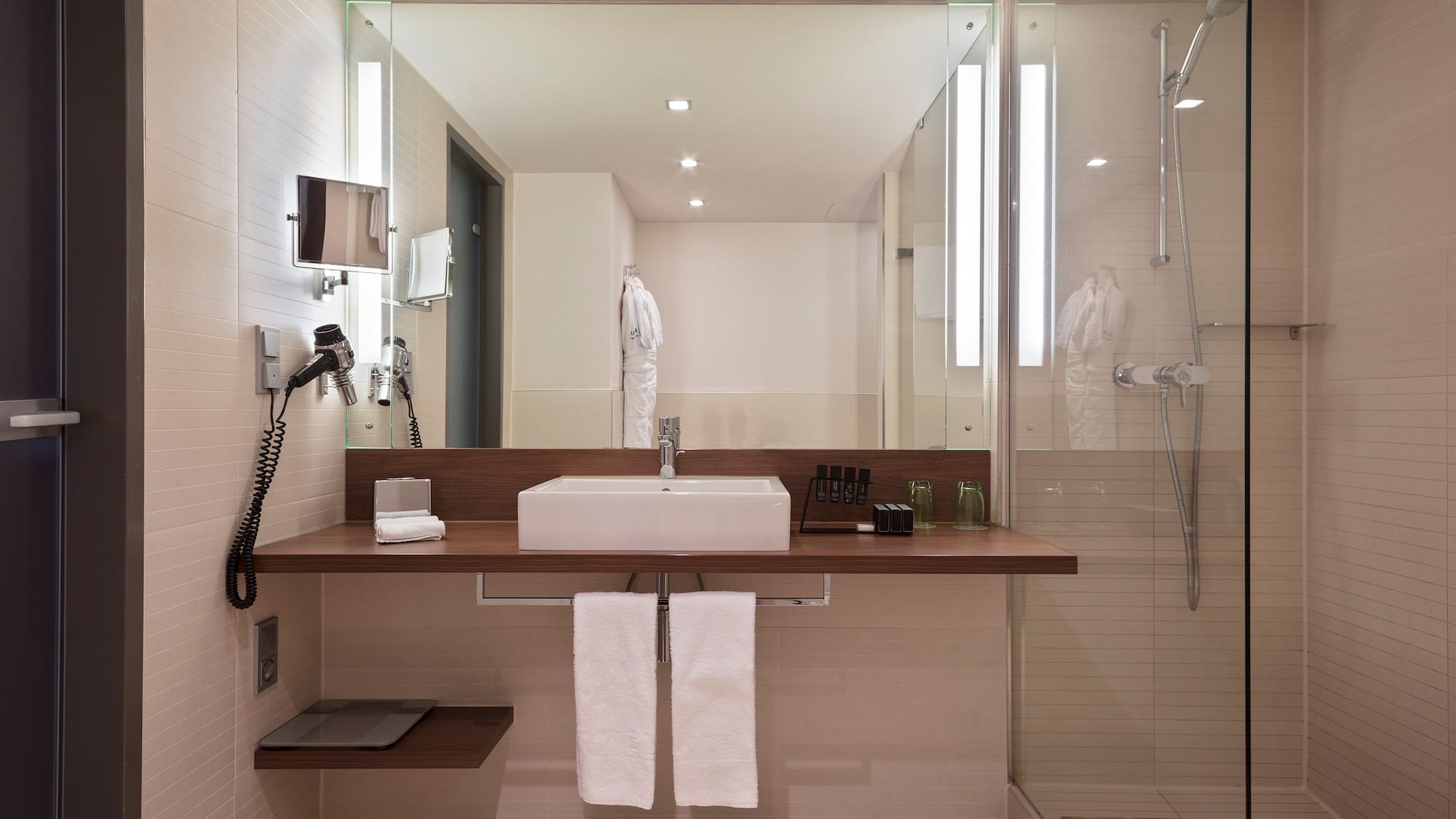 Modern hotel bathroom with white sink, wooden vanity, large mirror, and glass shower.