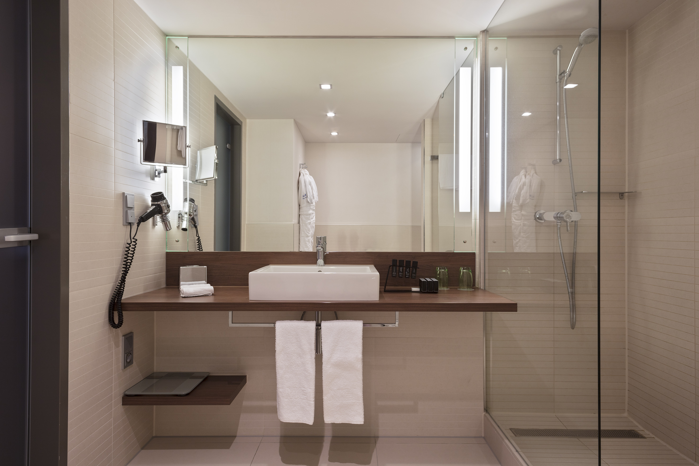 Modern hotel bathroom with white sink, wooden vanity, large mirror, and glass shower.