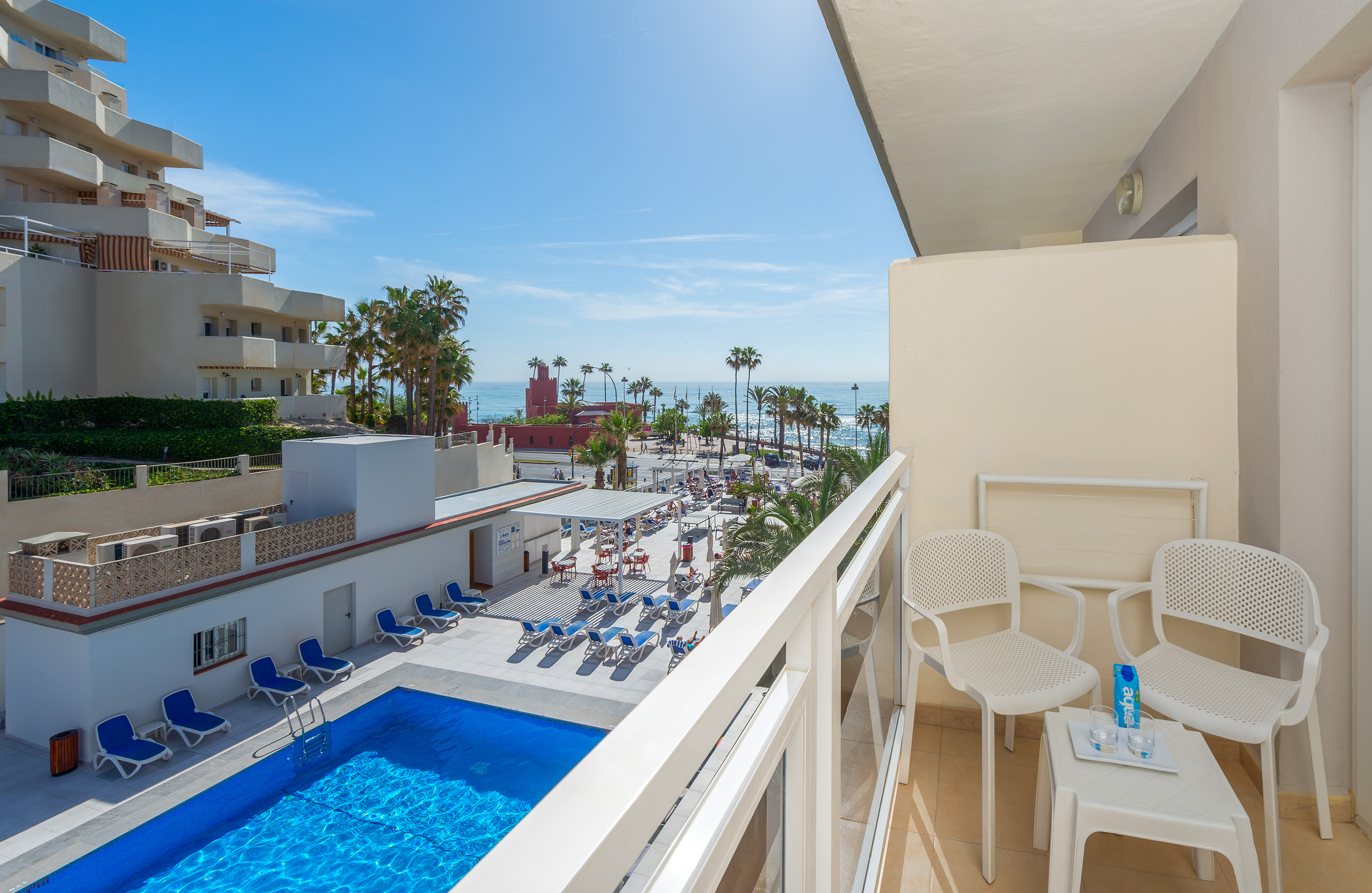 Hotel balcony view of sparkling pool, palm trees, and sunny oceanfront.