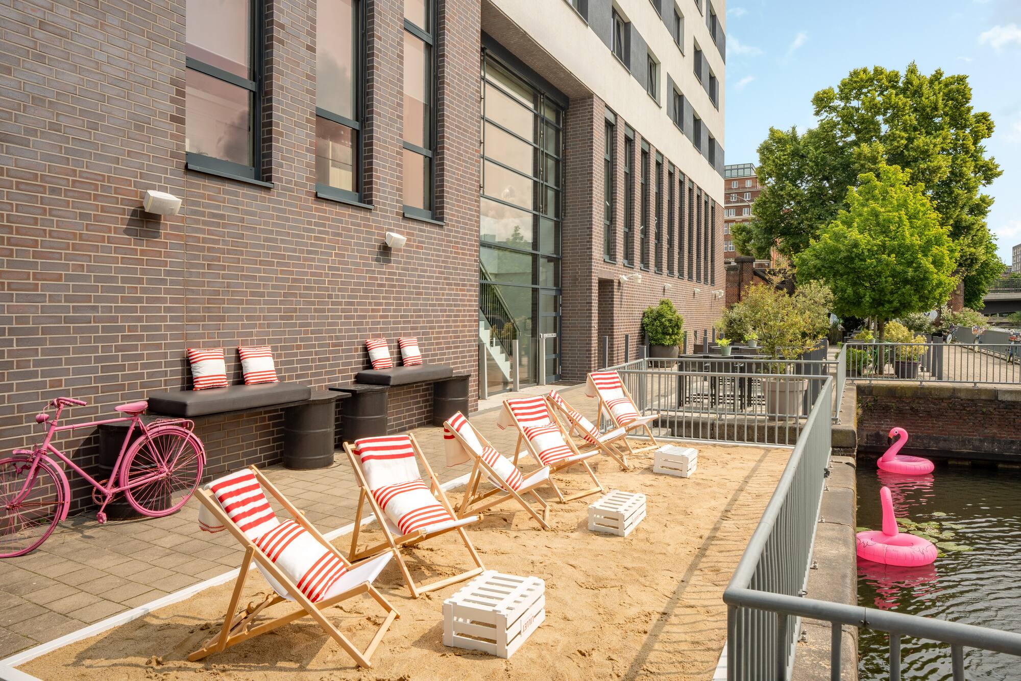 Urban sandy patio with beach chairs, pink bike, canal, modern building.