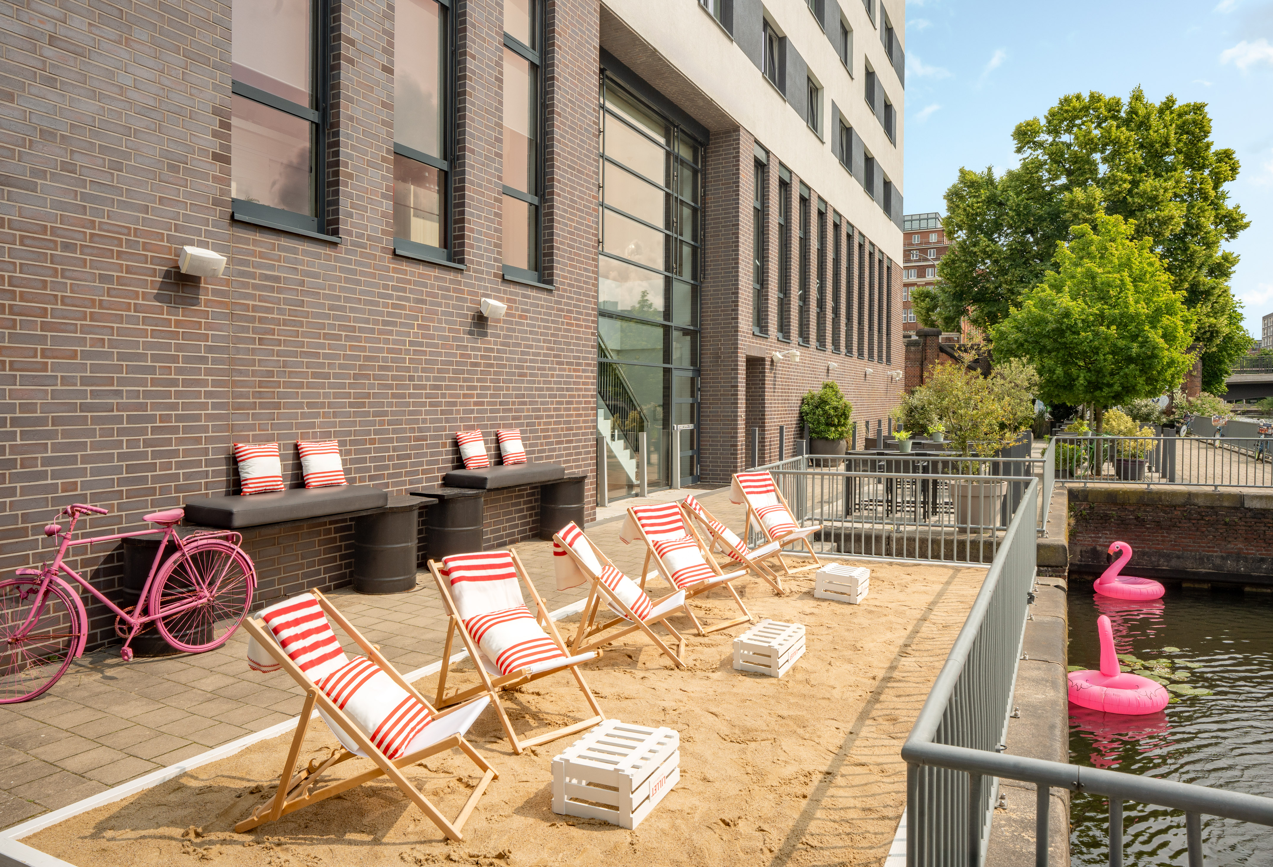 Urban sandy patio with beach chairs, pink bike, canal, modern building.