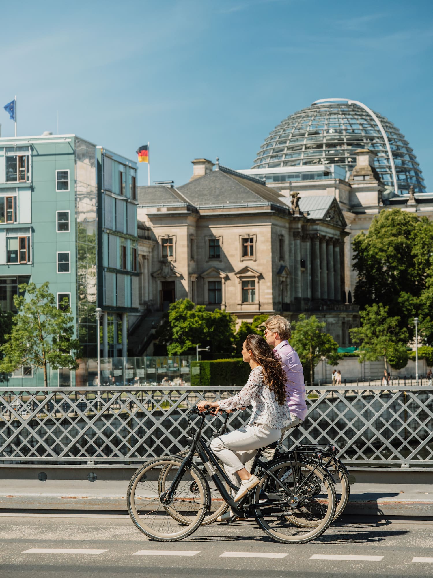 Couple biking on a bridge in Berlin with the Reichstag building dome and flag.