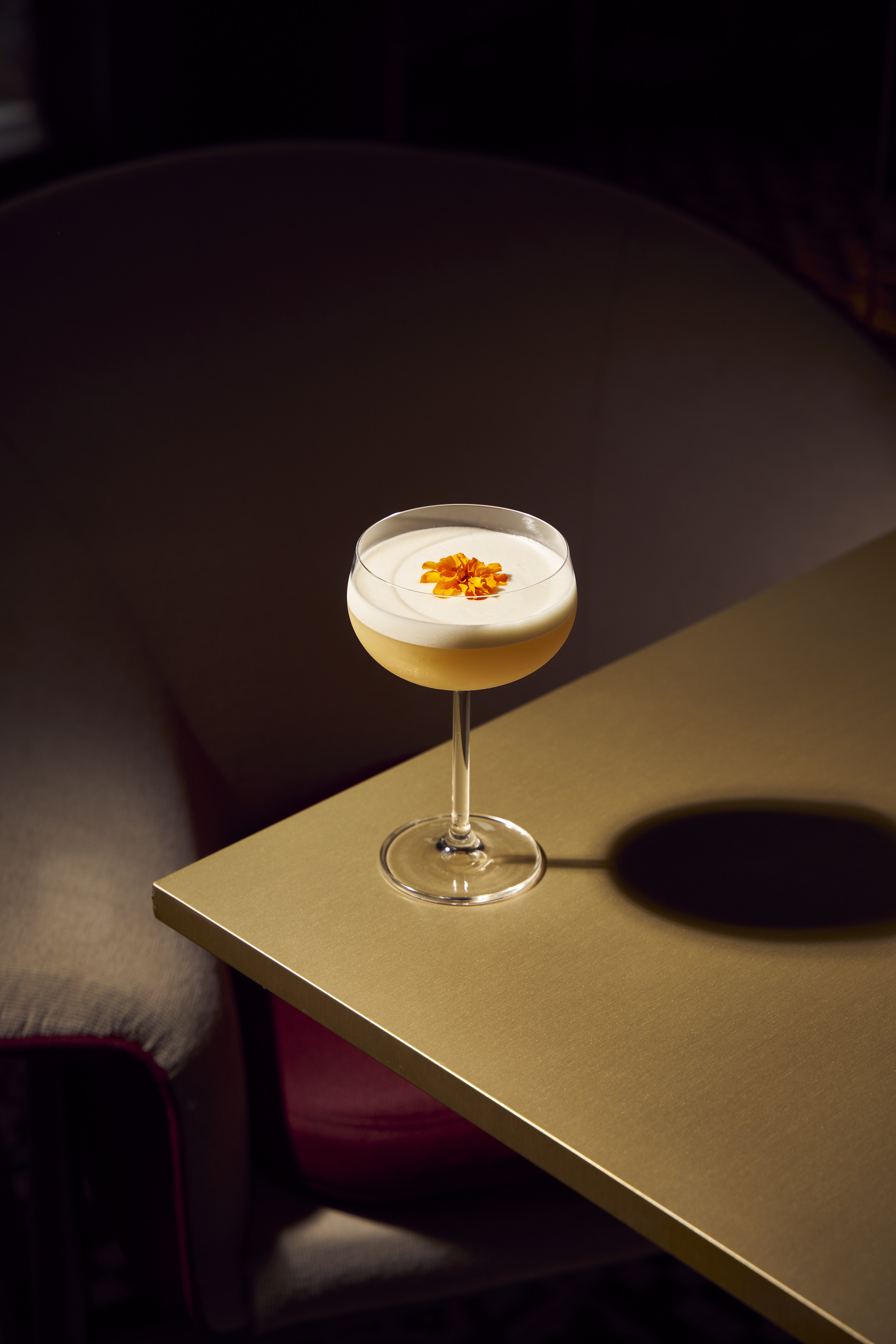 Yellow foamy cocktail with orange flower garnish in coupe glass on golden table.