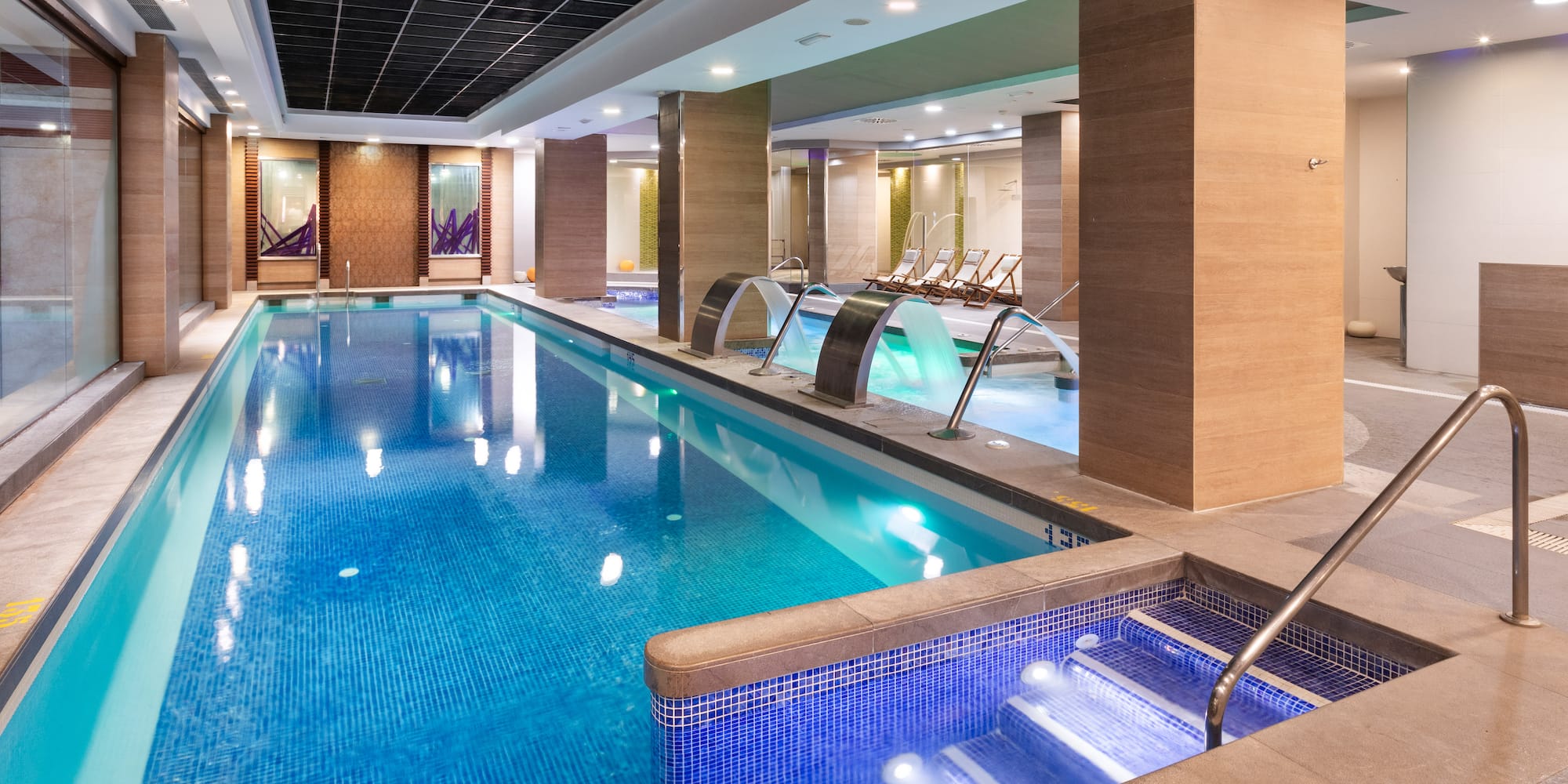 Modern indoor spa pool with blue mosaic tiles, relaxing water jets, and lounge chairs.