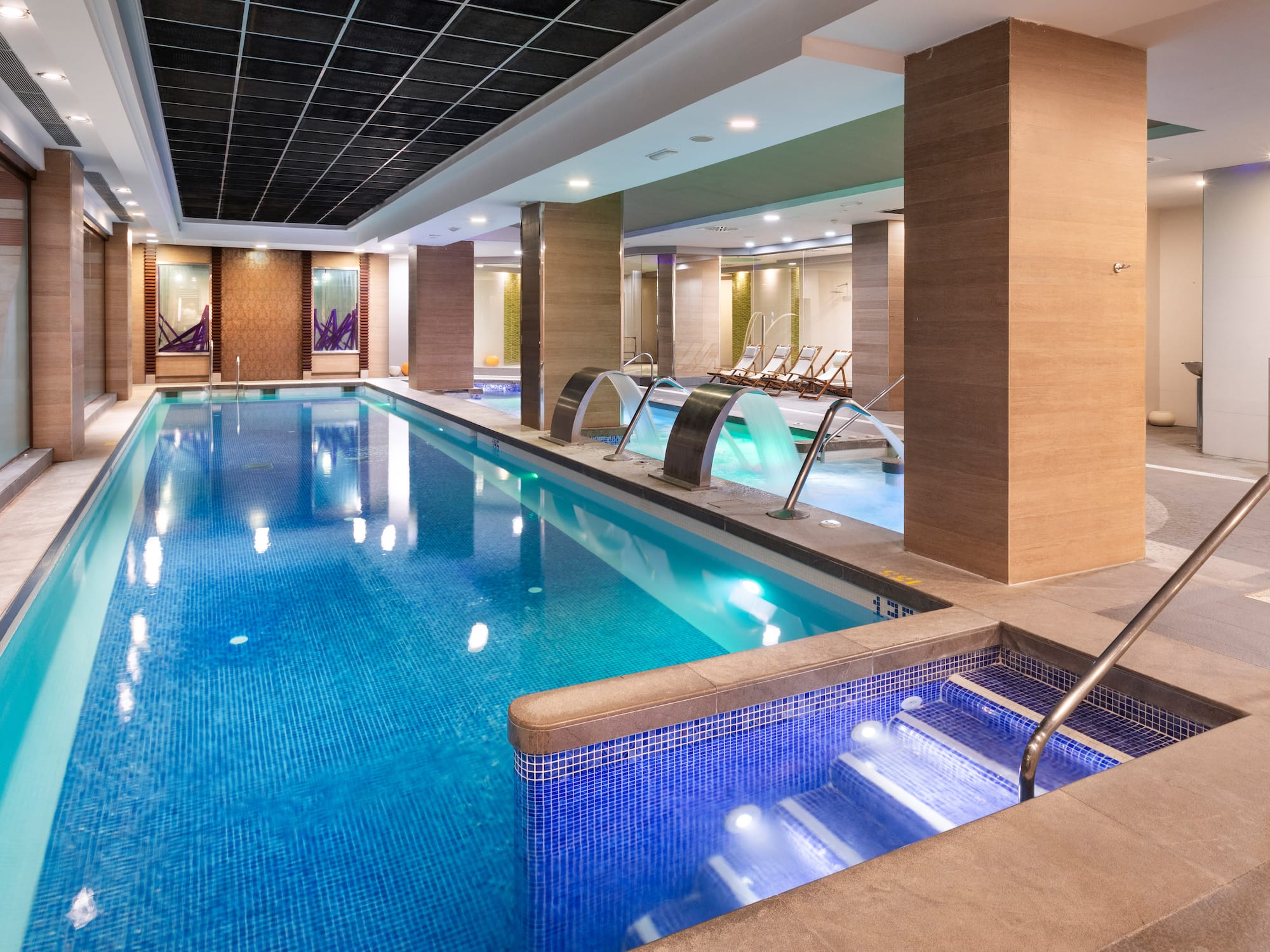 Indoor hotel swimming pool with blue tiles, water features, and spa loungers.