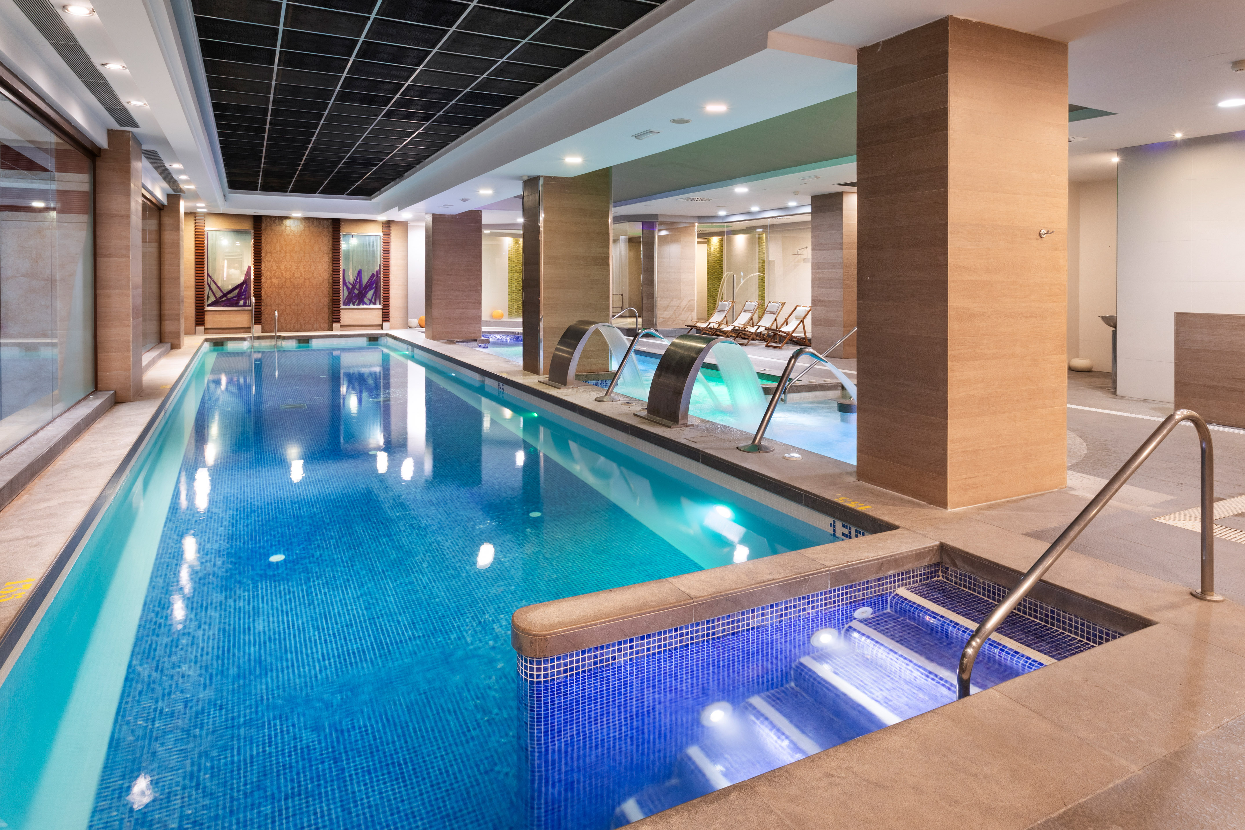 Indoor hotel swimming pool with blue tiles, water features, and spa loungers.