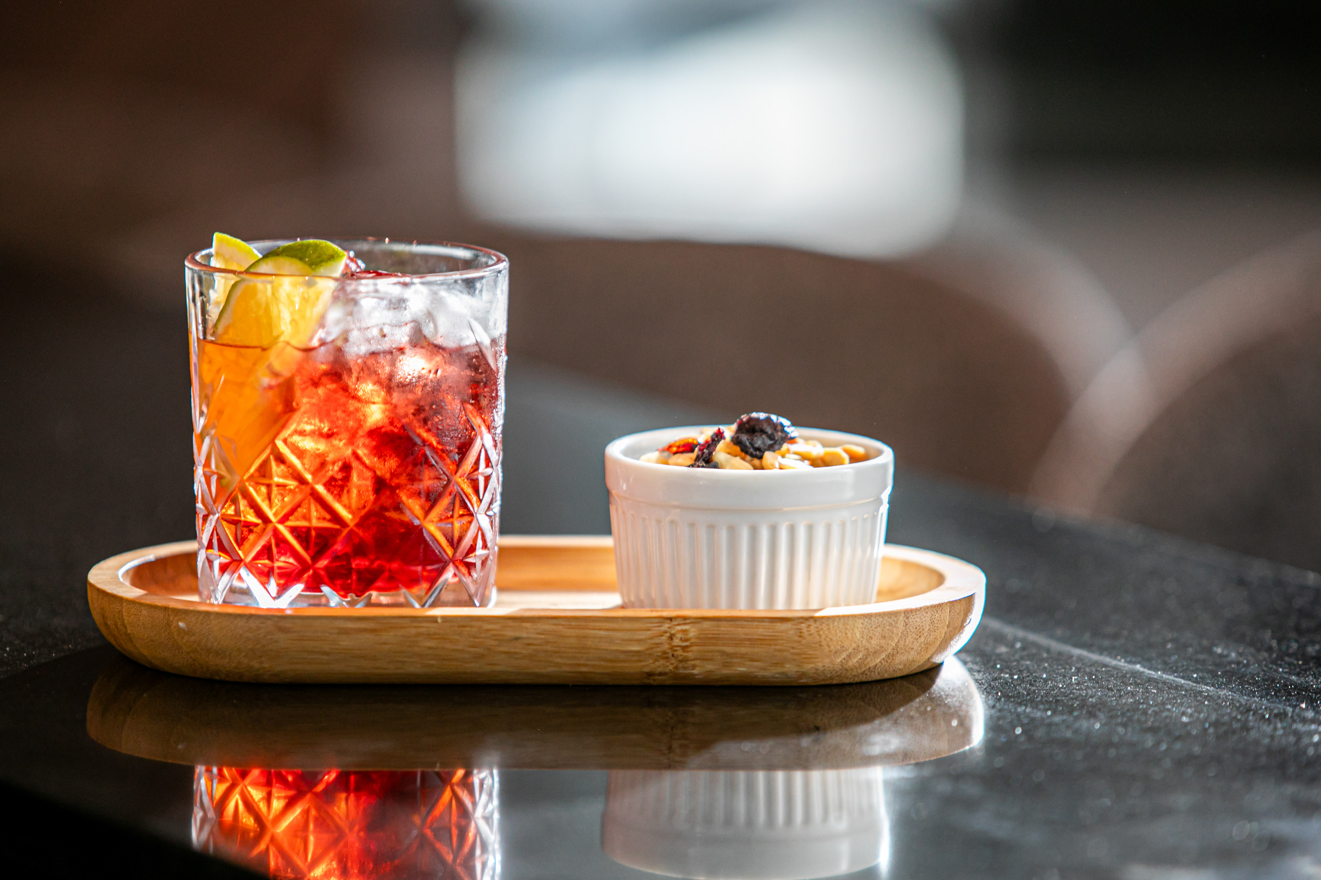 Red cocktail with orange, lime, and ice, with nuts on a wooden tray.