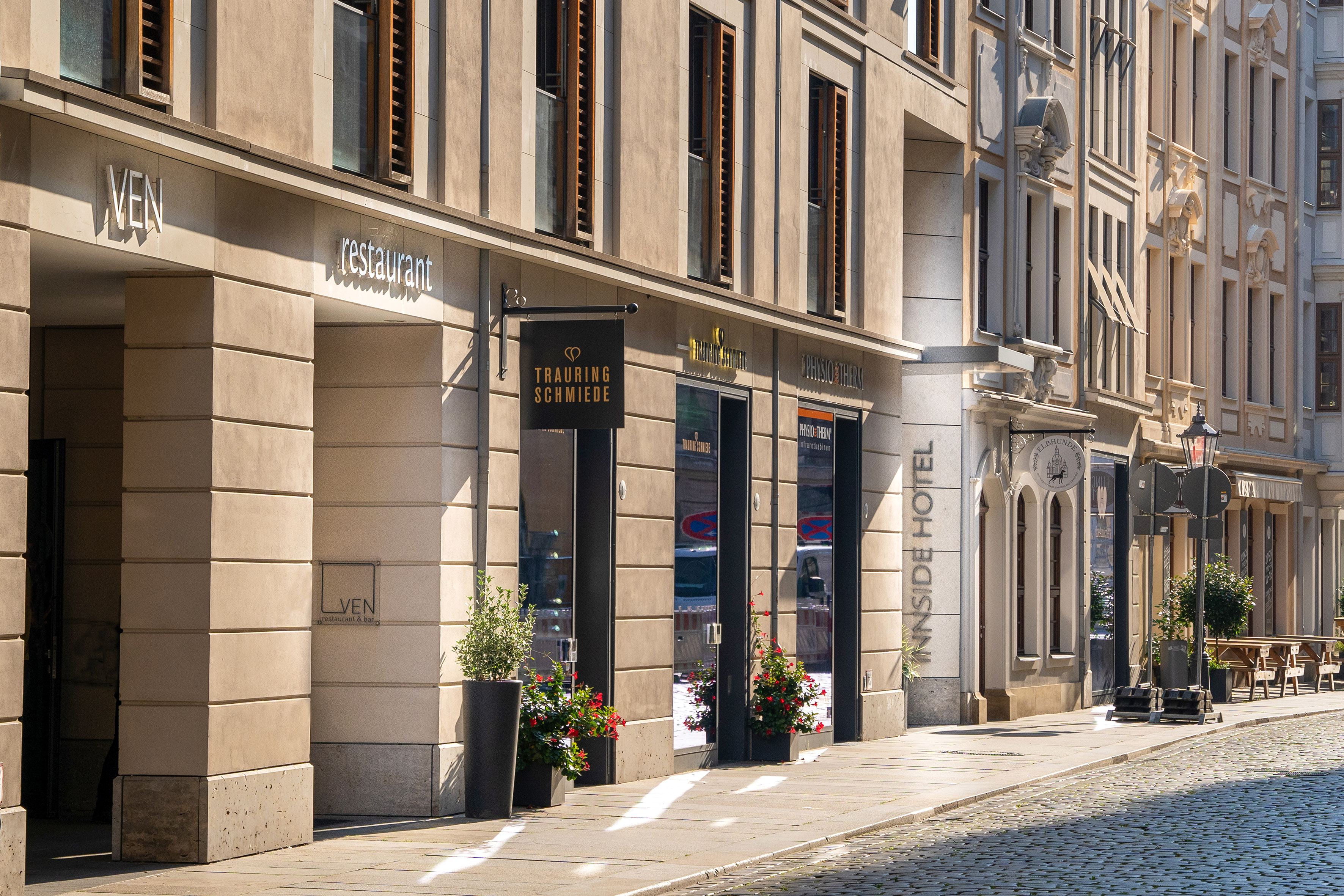 Sunny urban street with VEN restaurant, jewelry shop, and Innside Hotel storefronts.