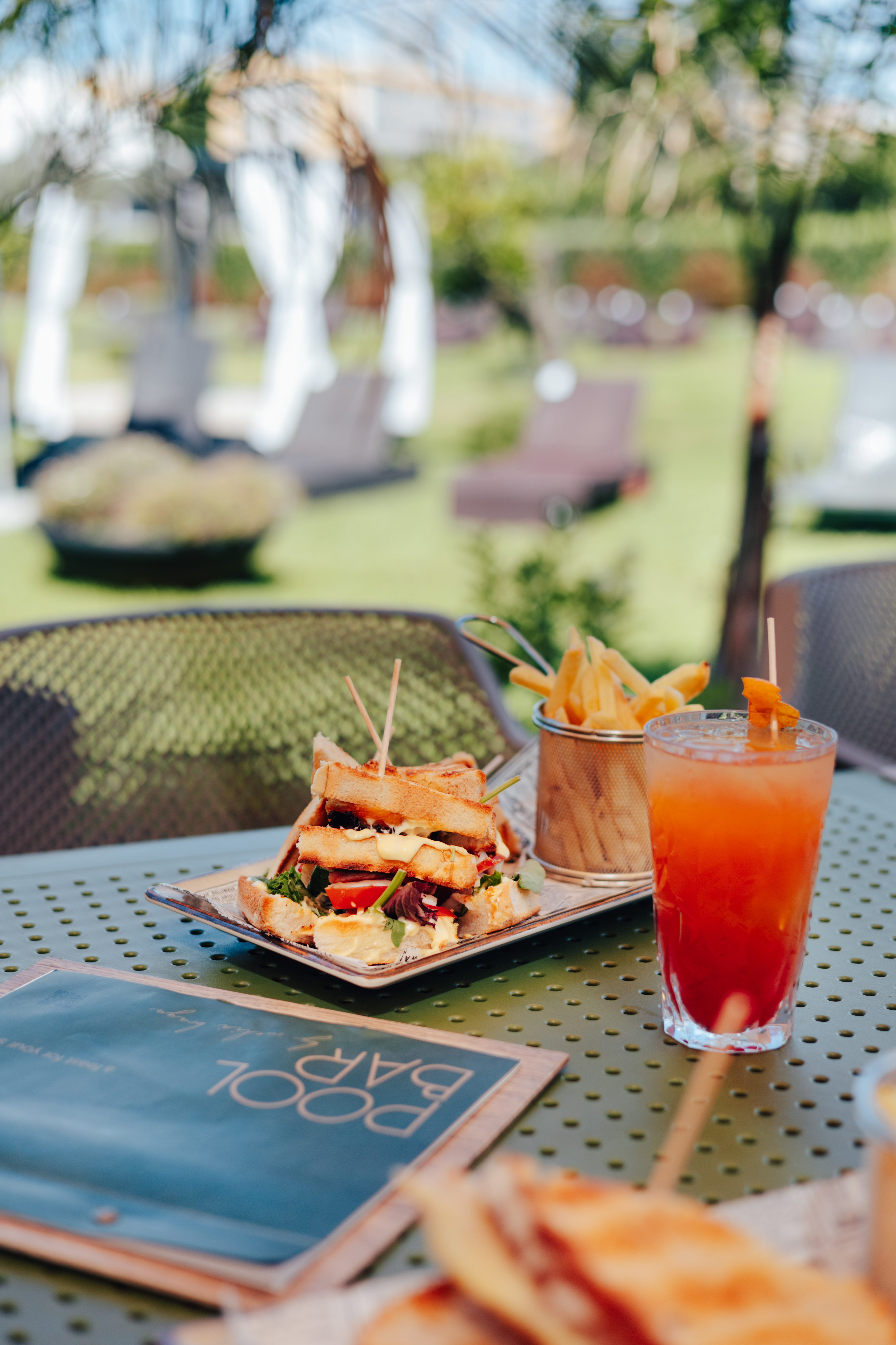 Club sandwich, fries, and cocktail on a table at an outdoor pool bar resort.