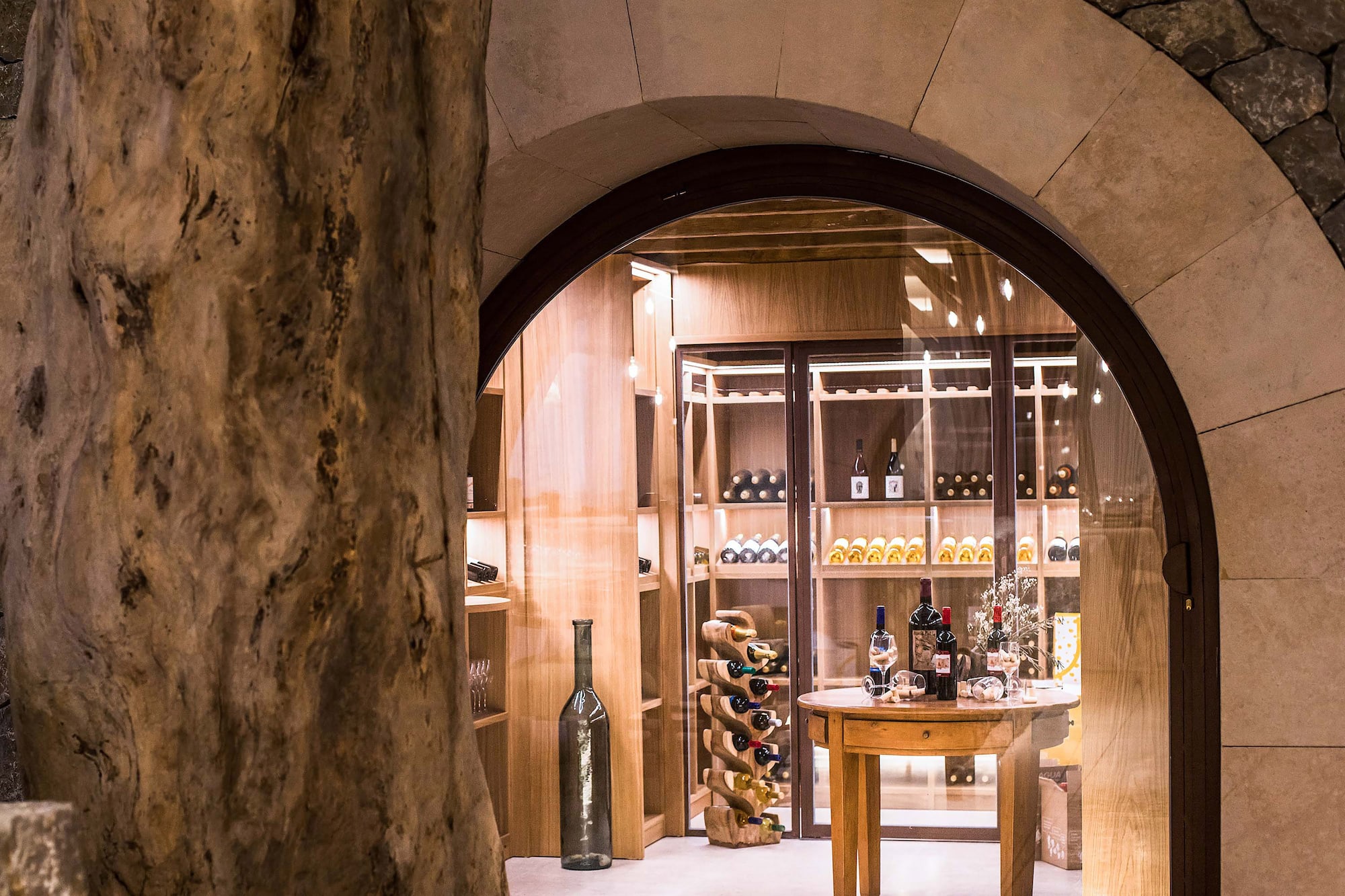 Rustic wine cellar with bottles, wooden racks, and arched stone entrance.