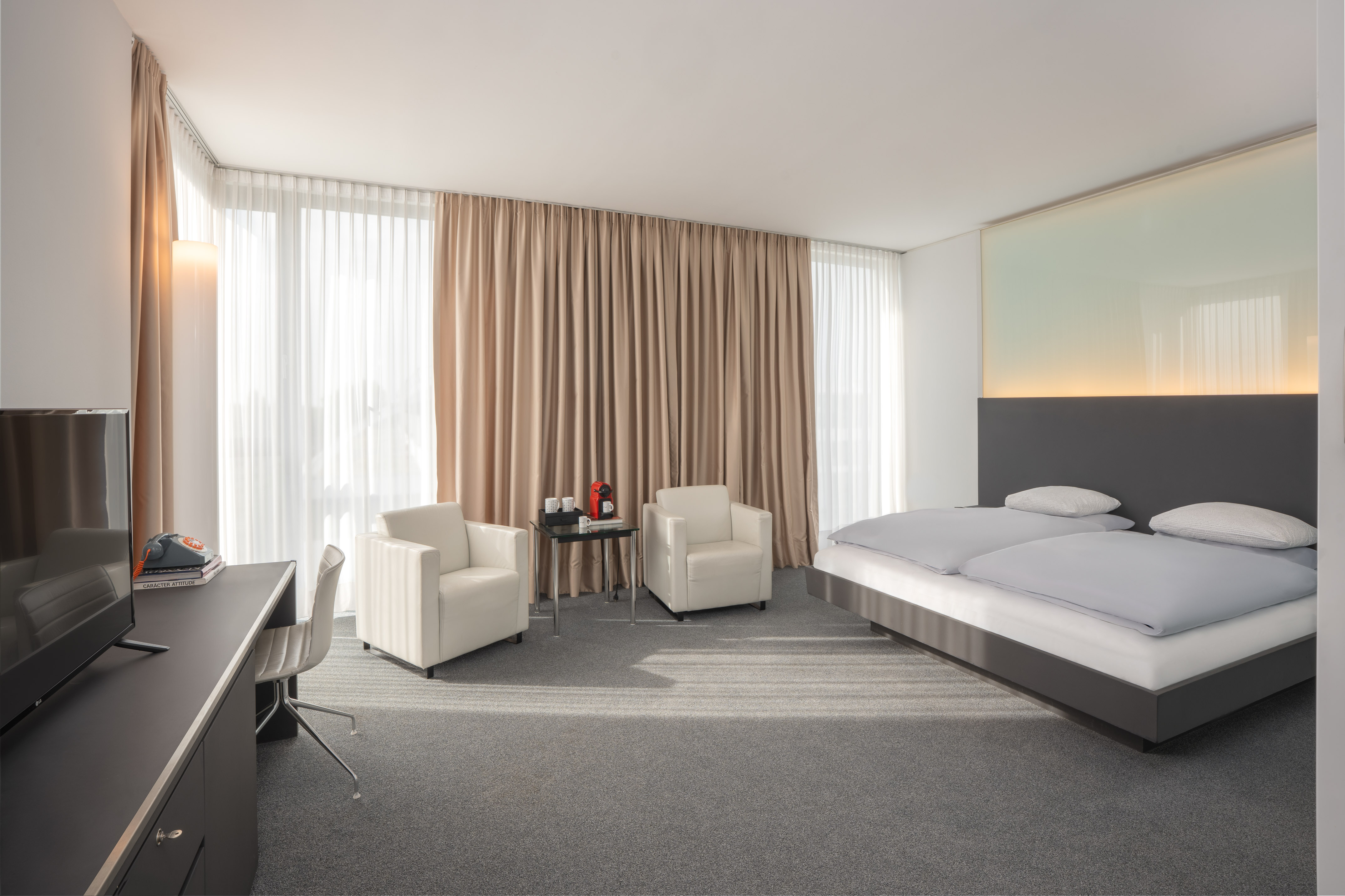 Contemporary hotel bedroom with king bed, armchairs, desk, and bright windows.