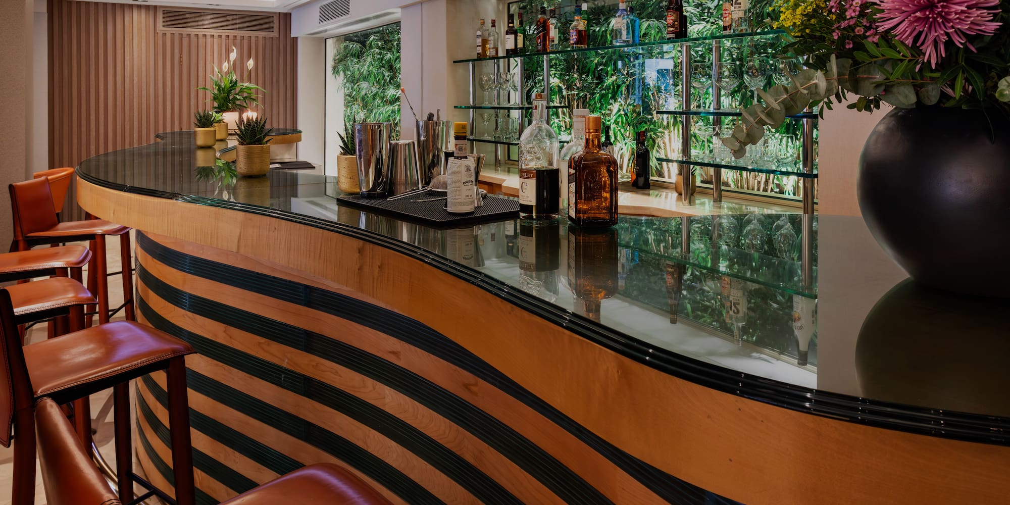 Elegant bar with striped wooden counter, leather stools, liquor, and green backdrop.