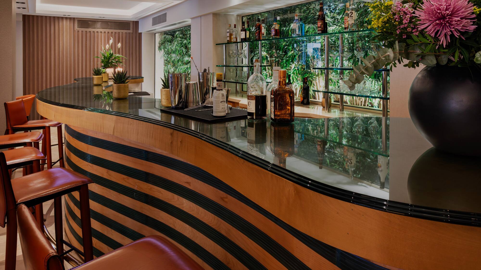 Elegant bar with striped wooden counter, leather stools, liquor, and green backdrop.