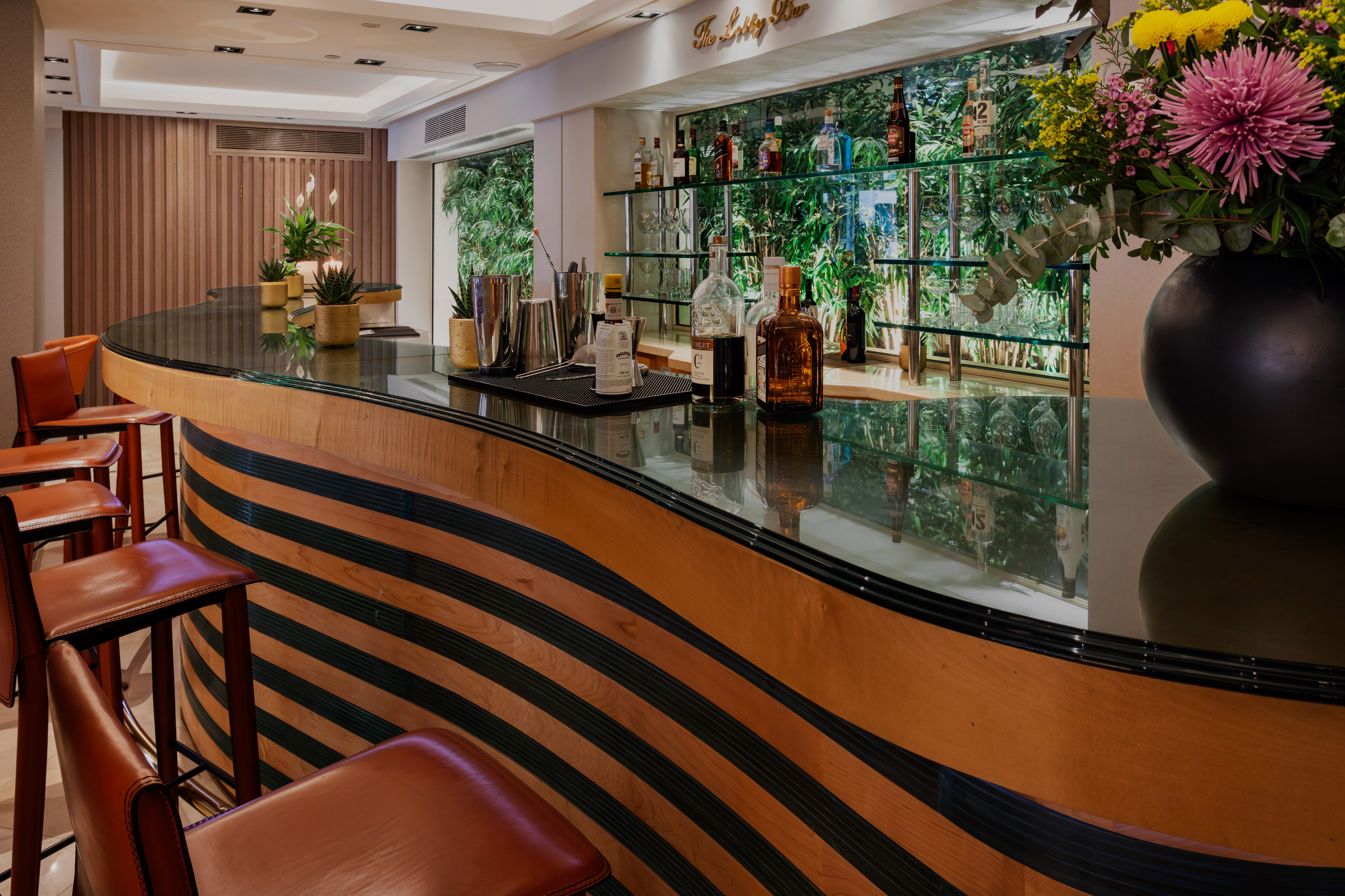 Elegant bar with striped wooden counter, leather stools, liquor, and green backdrop.