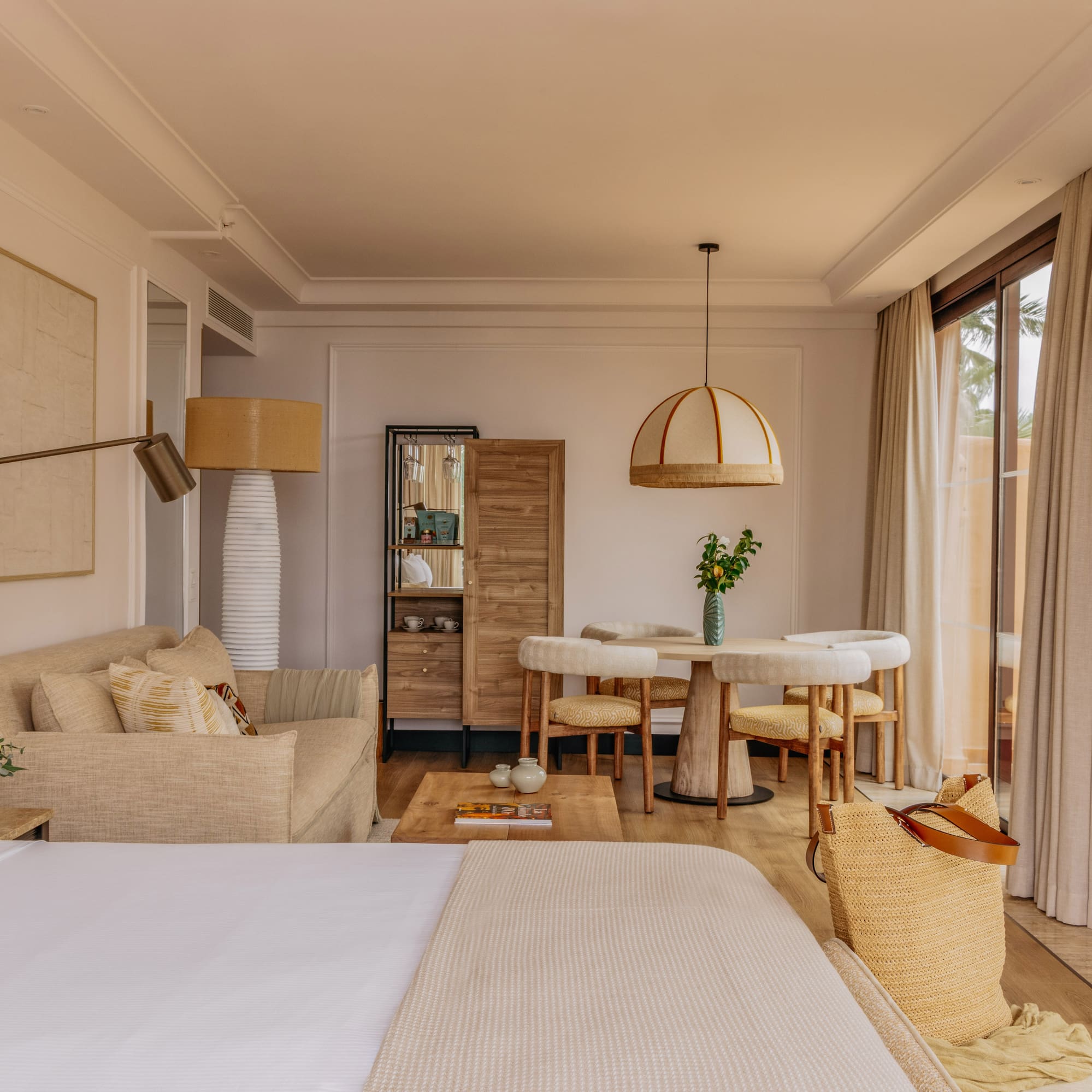 Modern coastal hotel suite with bed, sofa, dining set, and large window.