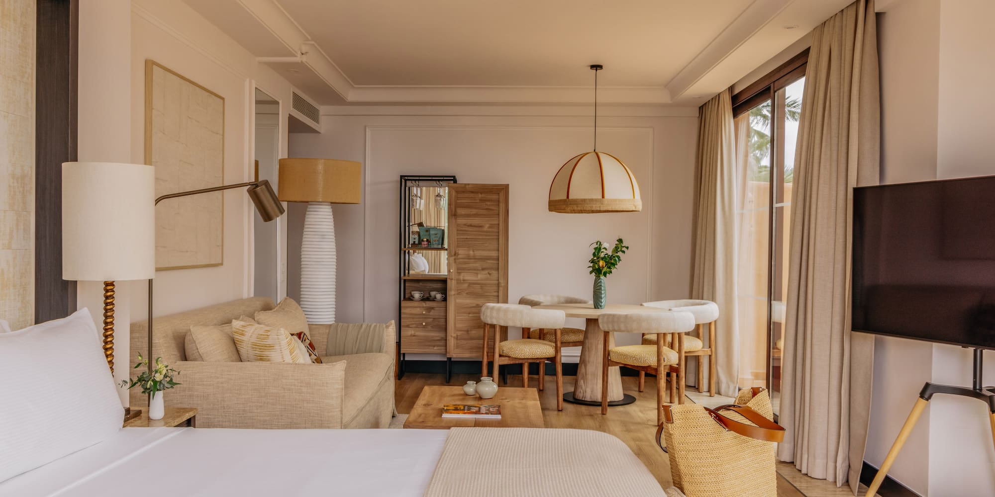 Modern coastal hotel suite with bed, sofa, dining set, and large window.