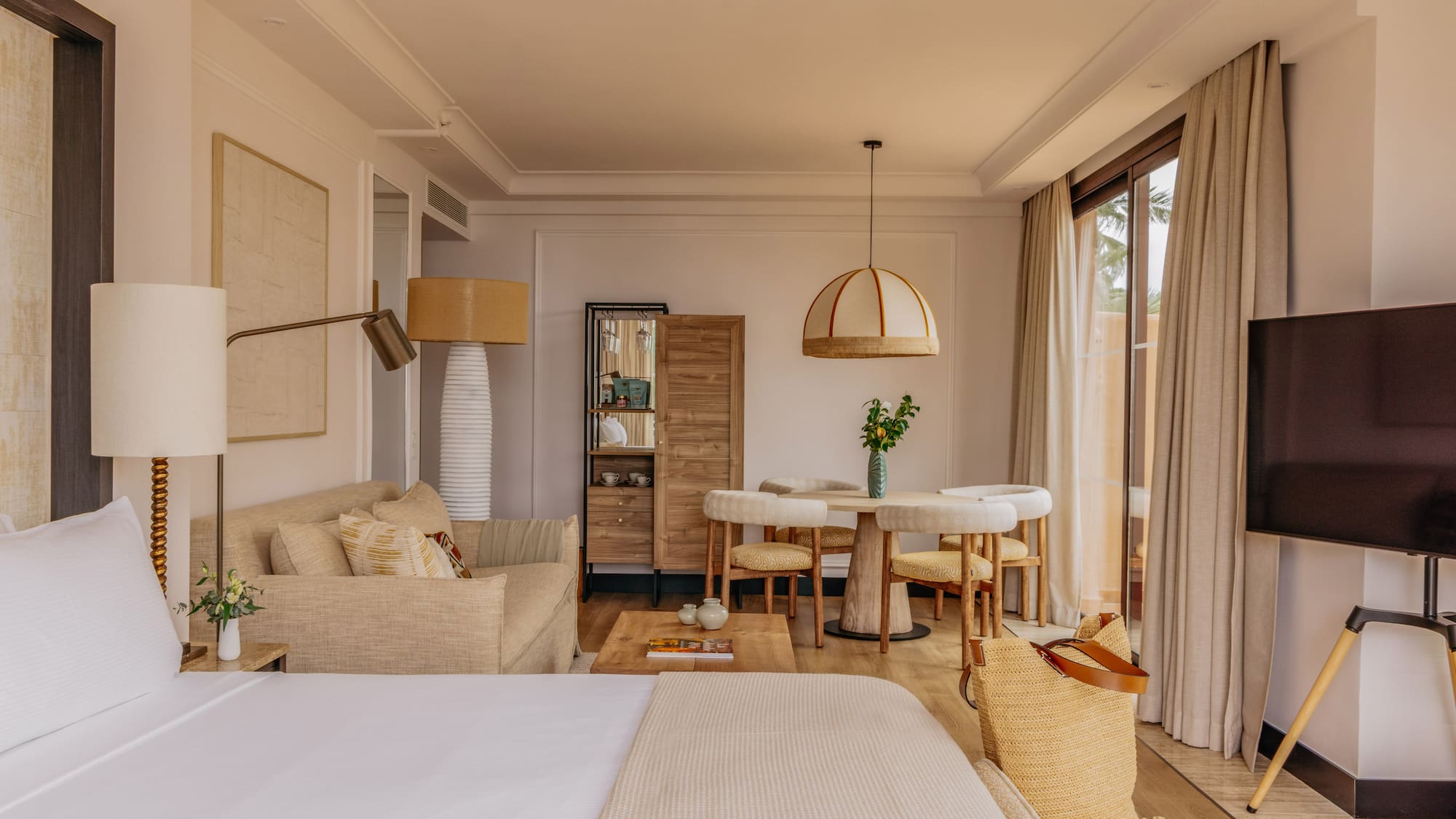 Modern coastal hotel suite with bed, sofa, dining set, and large window.