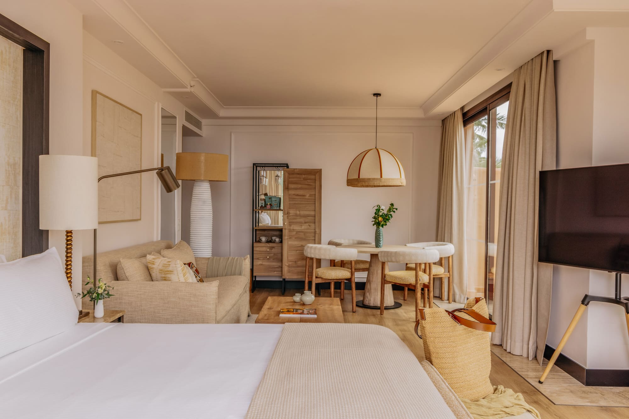 Modern coastal hotel suite with bed, sofa, dining set, and large window.