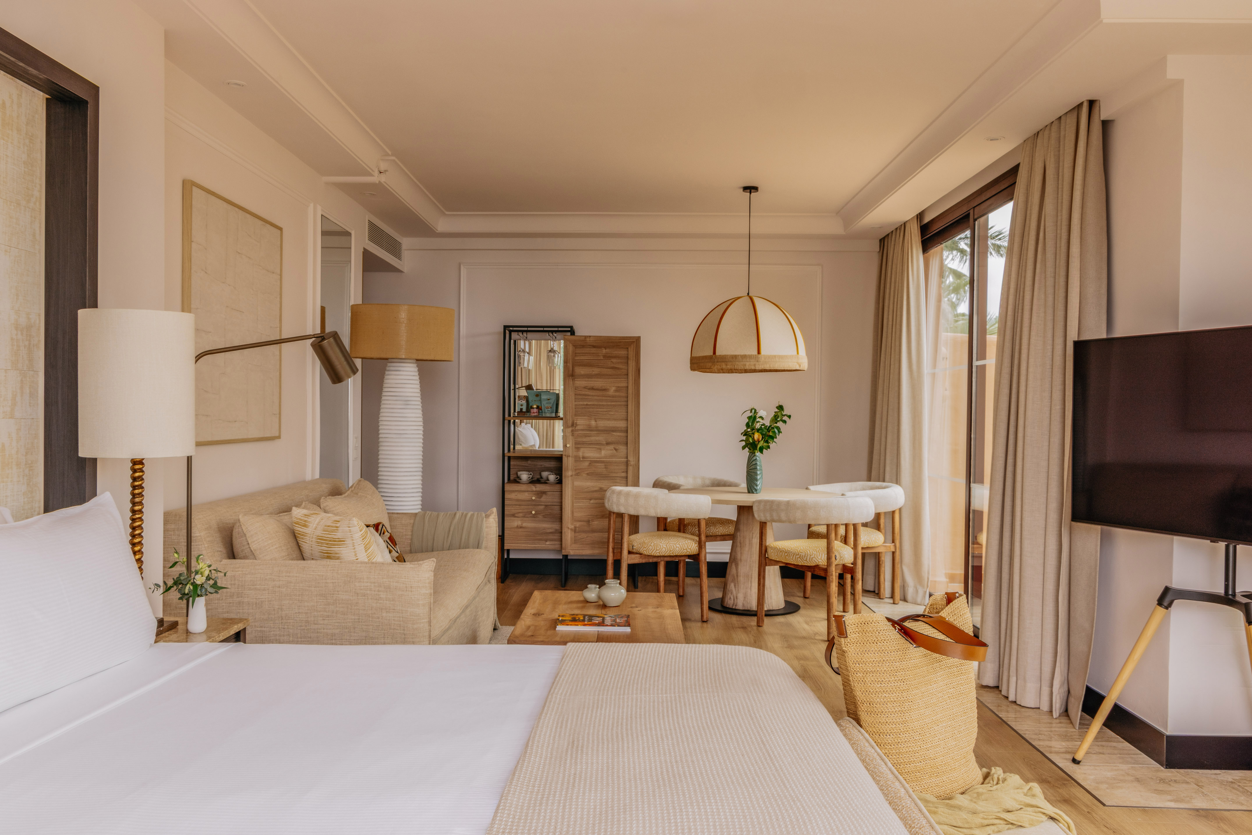 Modern coastal hotel suite with bed, sofa, dining set, and large window.