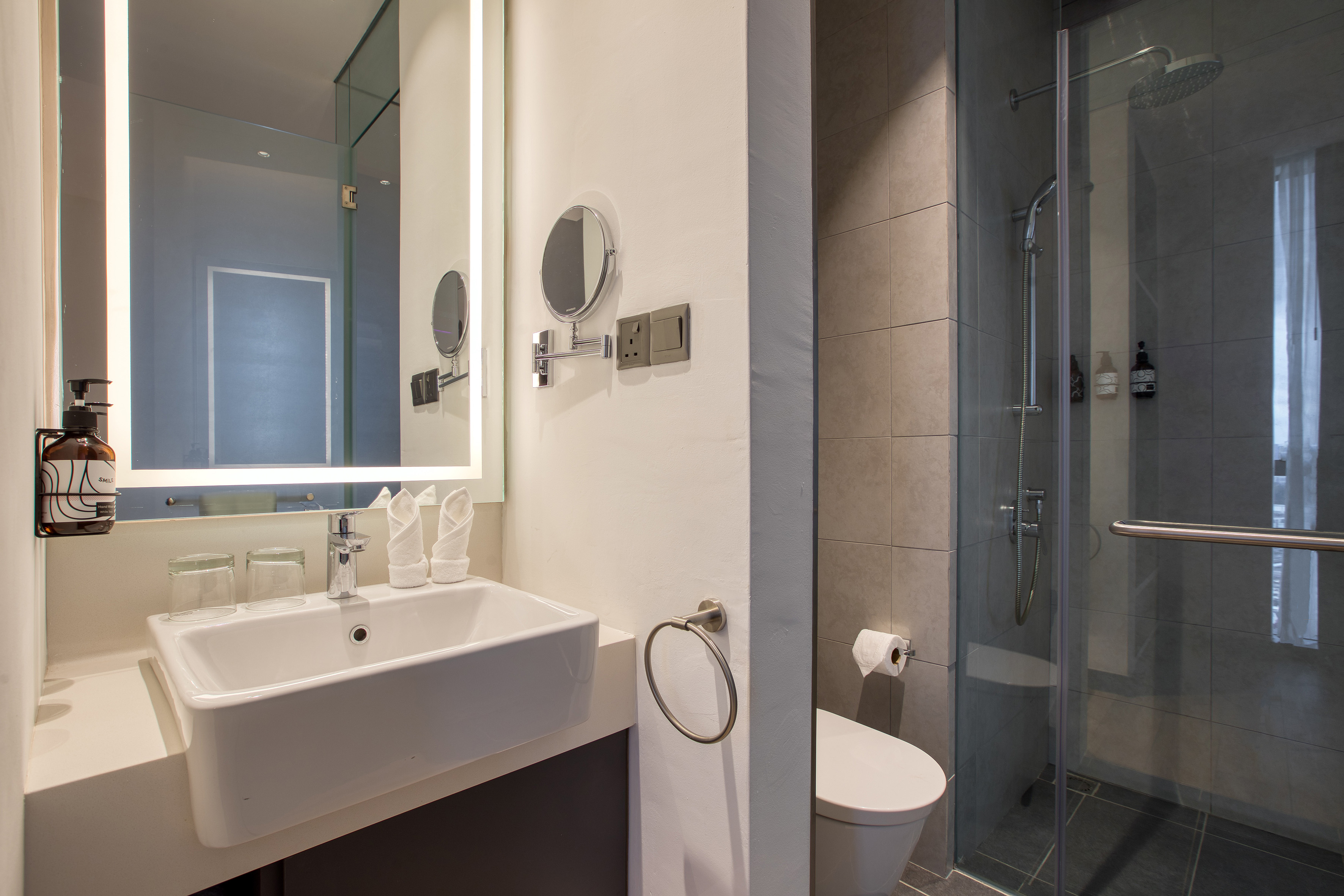 Modern hotel bathroom with illuminated mirror, white sink, glass shower, and toilet.