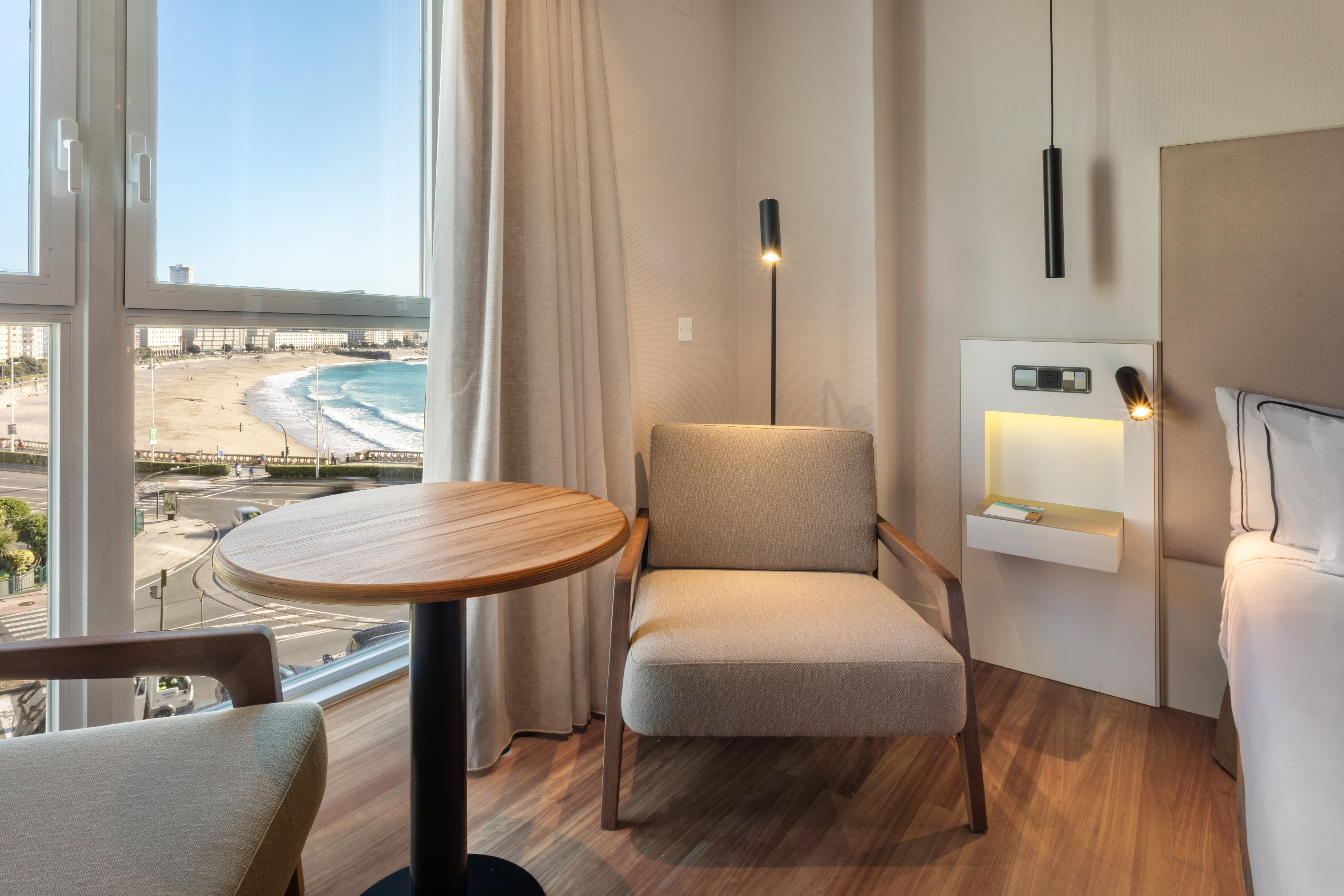 Modern hotel room overlooking a beach and ocean, with table and armchair.