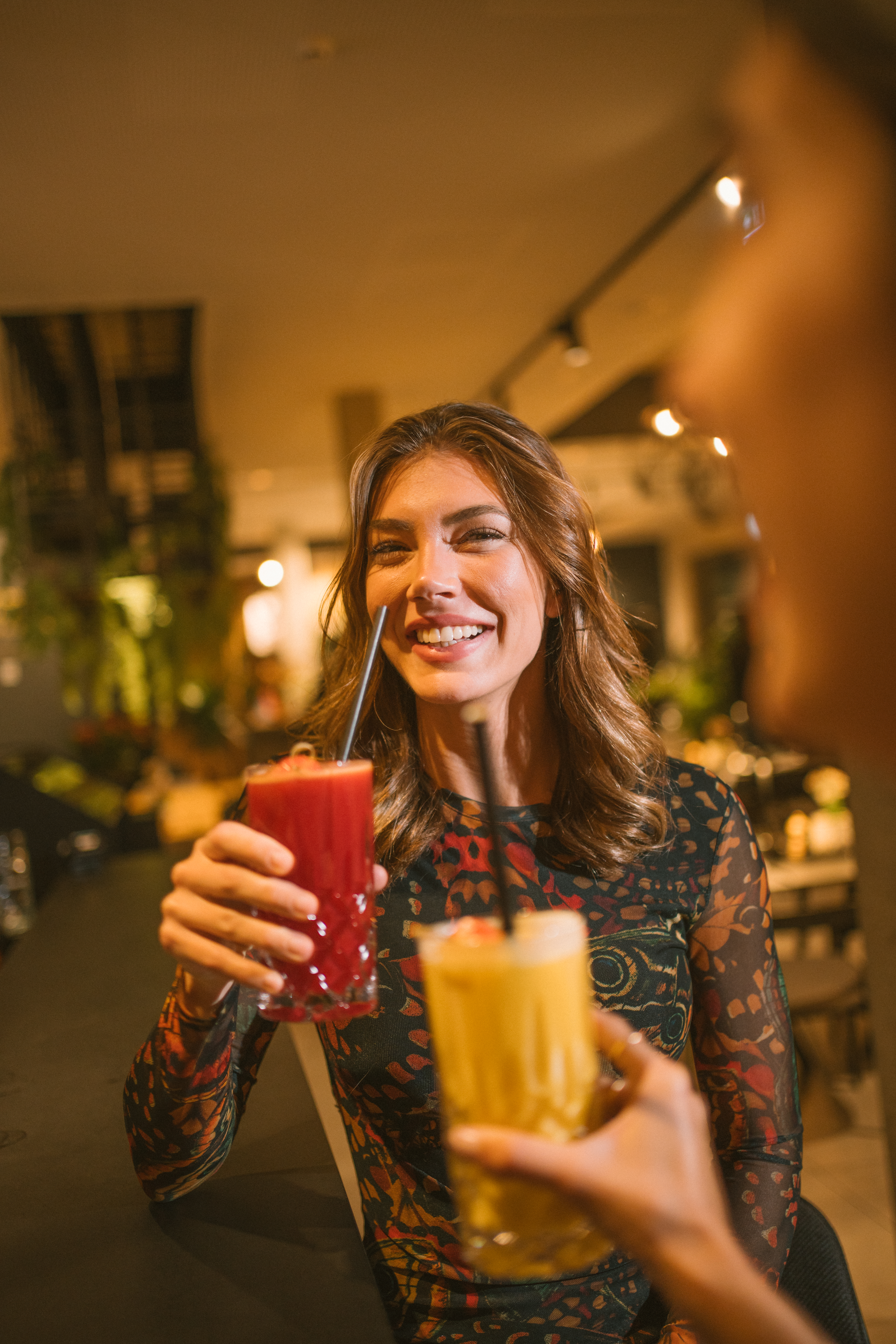 Smiling woman with a red cocktail clinking glasses with a friend at a bar.